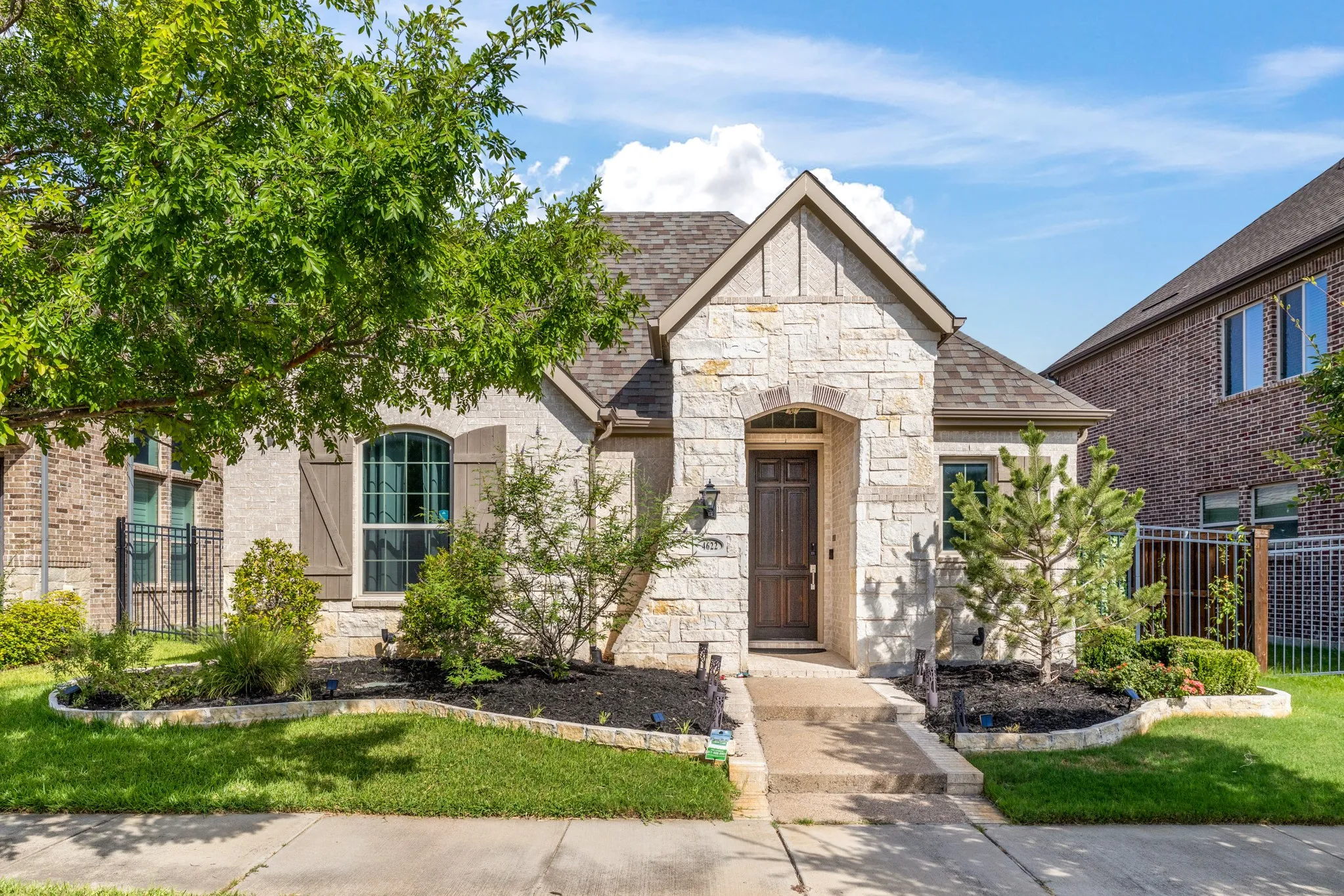 Elegant stone and brick exterior featuring a gabled roofline with arched entryway, complemented by manicured landscaping with mature trees, lush lawn, and defined planting beds