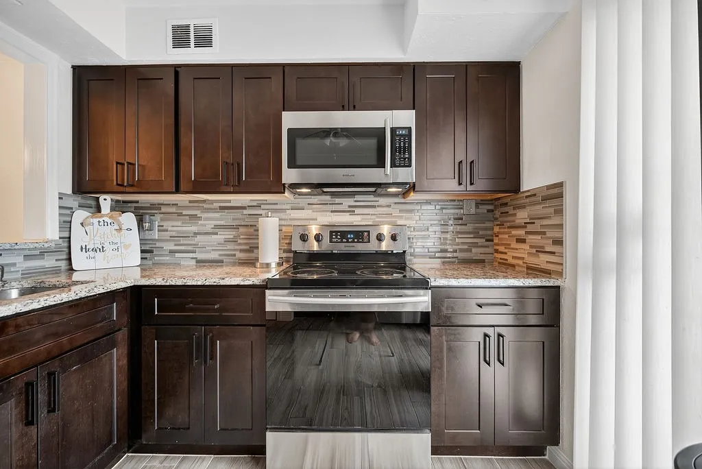 Modern kitchen featuring rich wood cabinetry, patterned tile backsplash, speckled countertops, stainless steel appliances, and wood-finish flooring