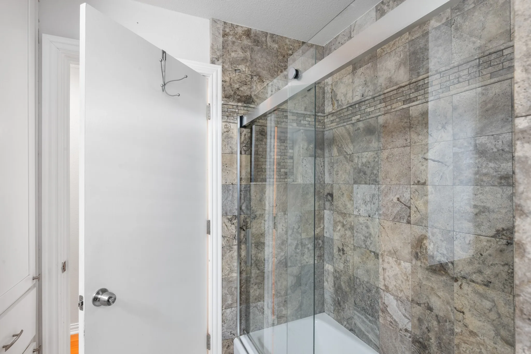 Modern bathroom featuring a frameless glass shower enclosure with brushed nickel hardware, natural stone tile surround in neutral earth tones, white subway tile accent wall, and a contemporary white door entry