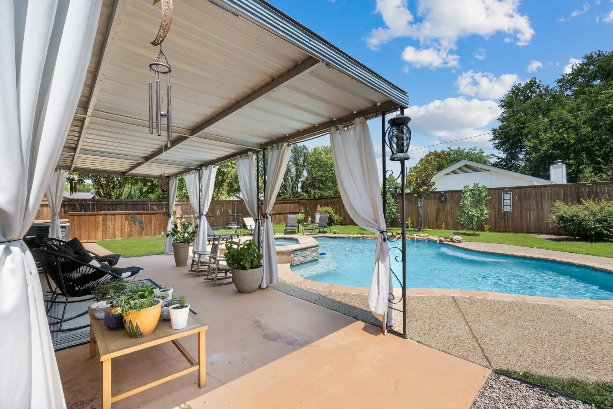 Resort-Style Backyard Oasis featuring an In-Ground Swimming Pool with crystal-clear waters, expansive concrete patio with integrated lounge seating, motorized pergola with retractable shade panels and sheer curtains, ambient outdoor lighting, and mature landscaping with privacy fencing