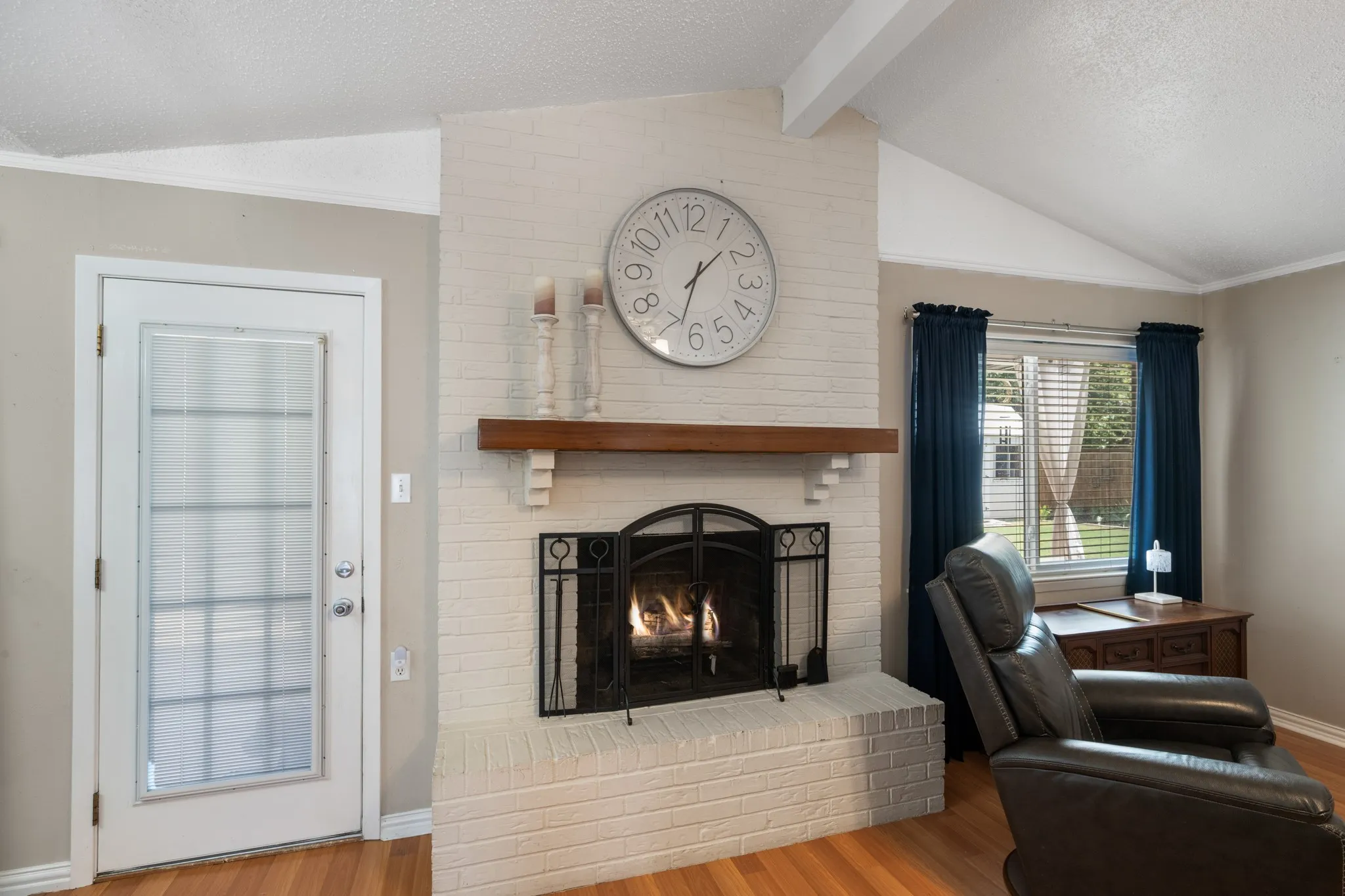 Bright living room showcasing a brick fireplace with black metal surround and mantel, hardwood flooring, large windows with dark drapery, vaulted ceilings, and a wall-mounted clock