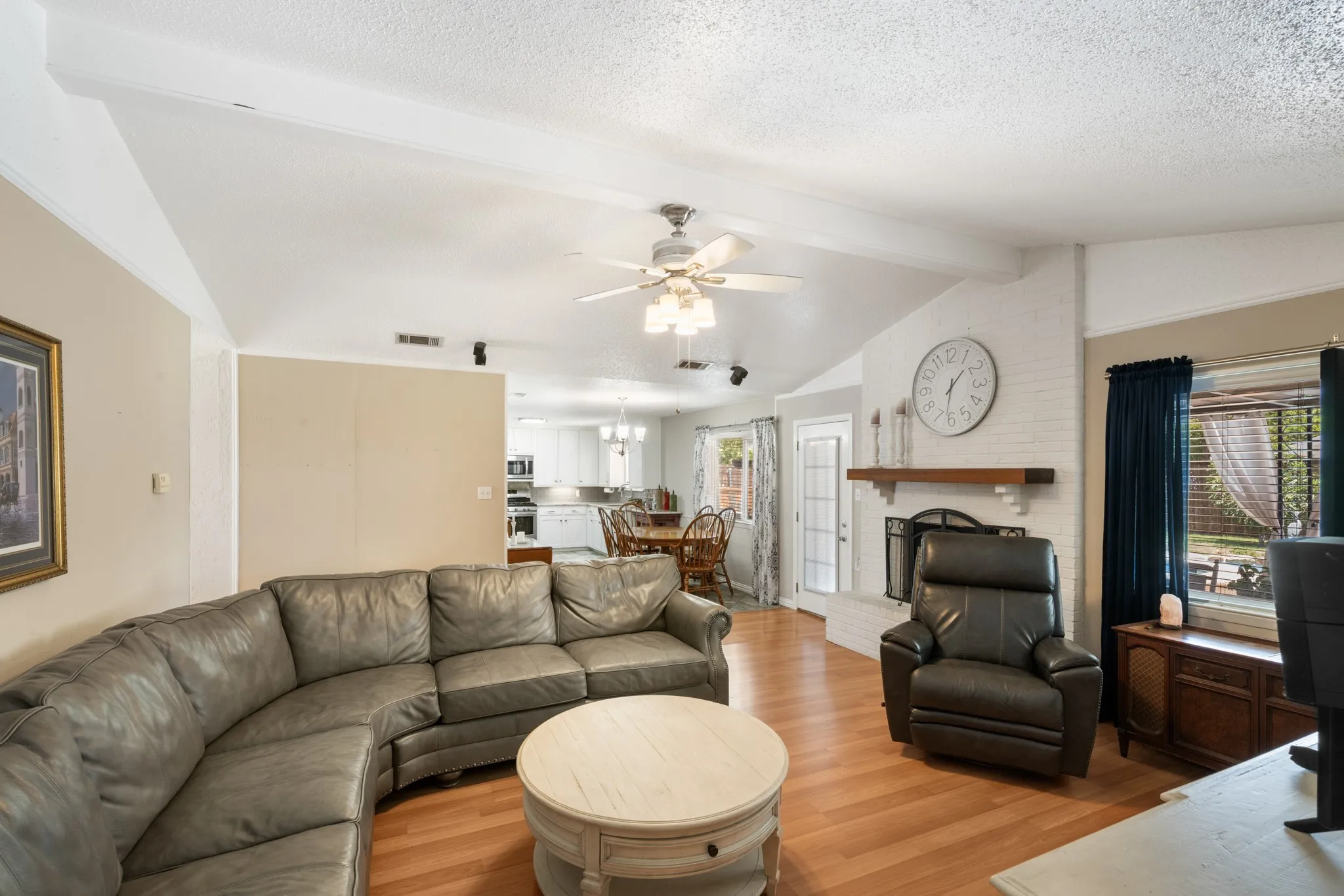Vaulted ceilings with exposed beams, wood-finish flooring, and a whitewashed brick fireplace with a wood mantel