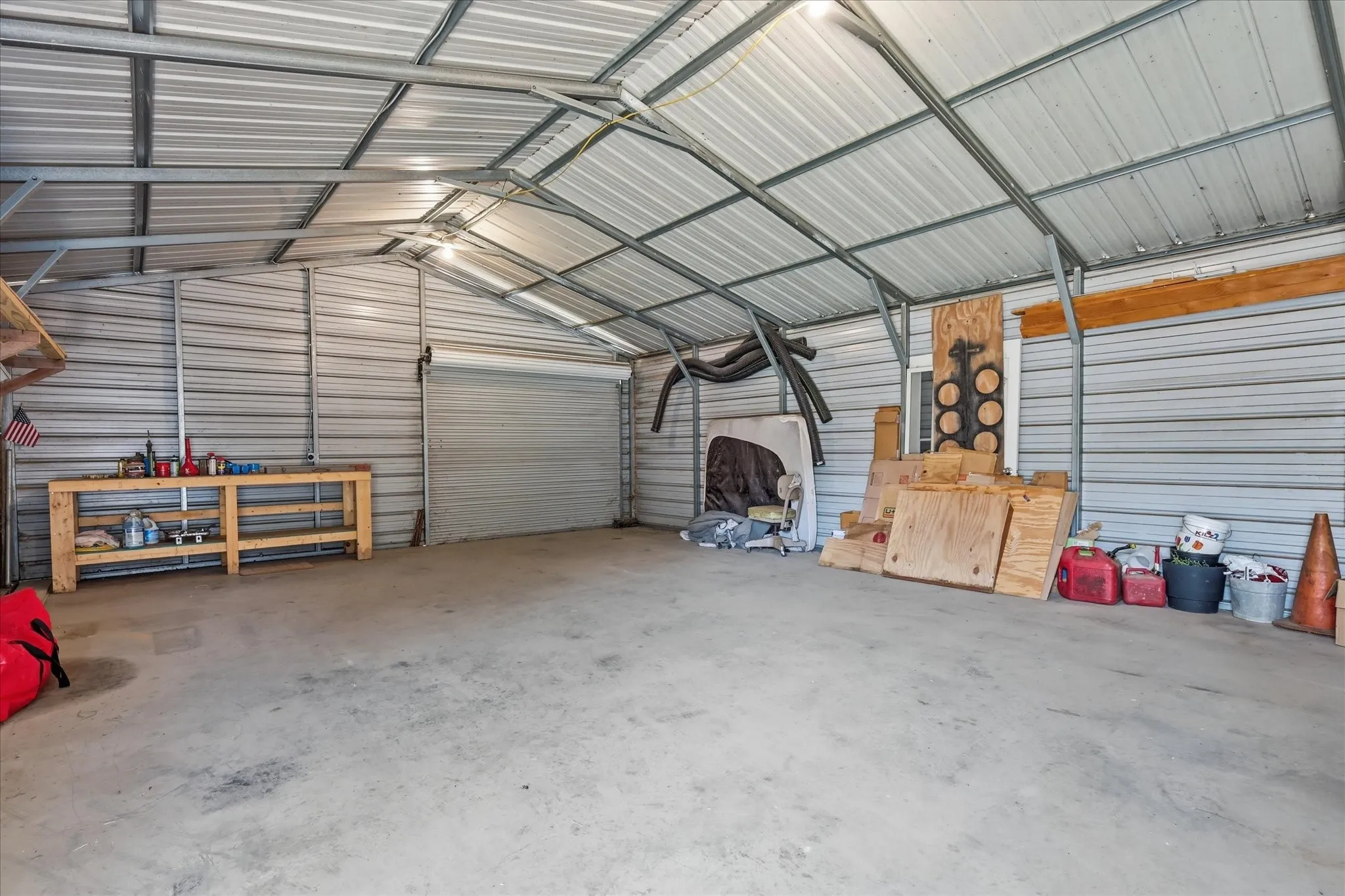 Spacious metal carport structure featuring a concrete floor, roll-up door, exposed metal beam ceiling, and integrated overhead lighting