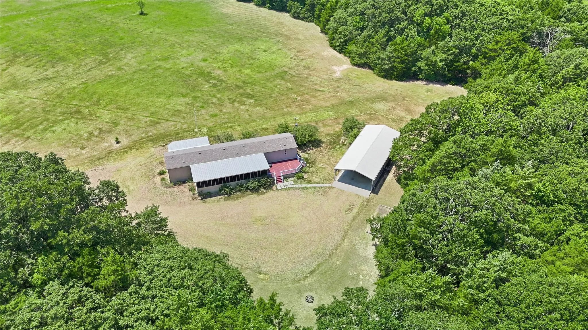 Expansive rural property featuring a primary residence with a screened porch and attached deck, a large detached metal carport structure, and surrounding mature trees and open fields