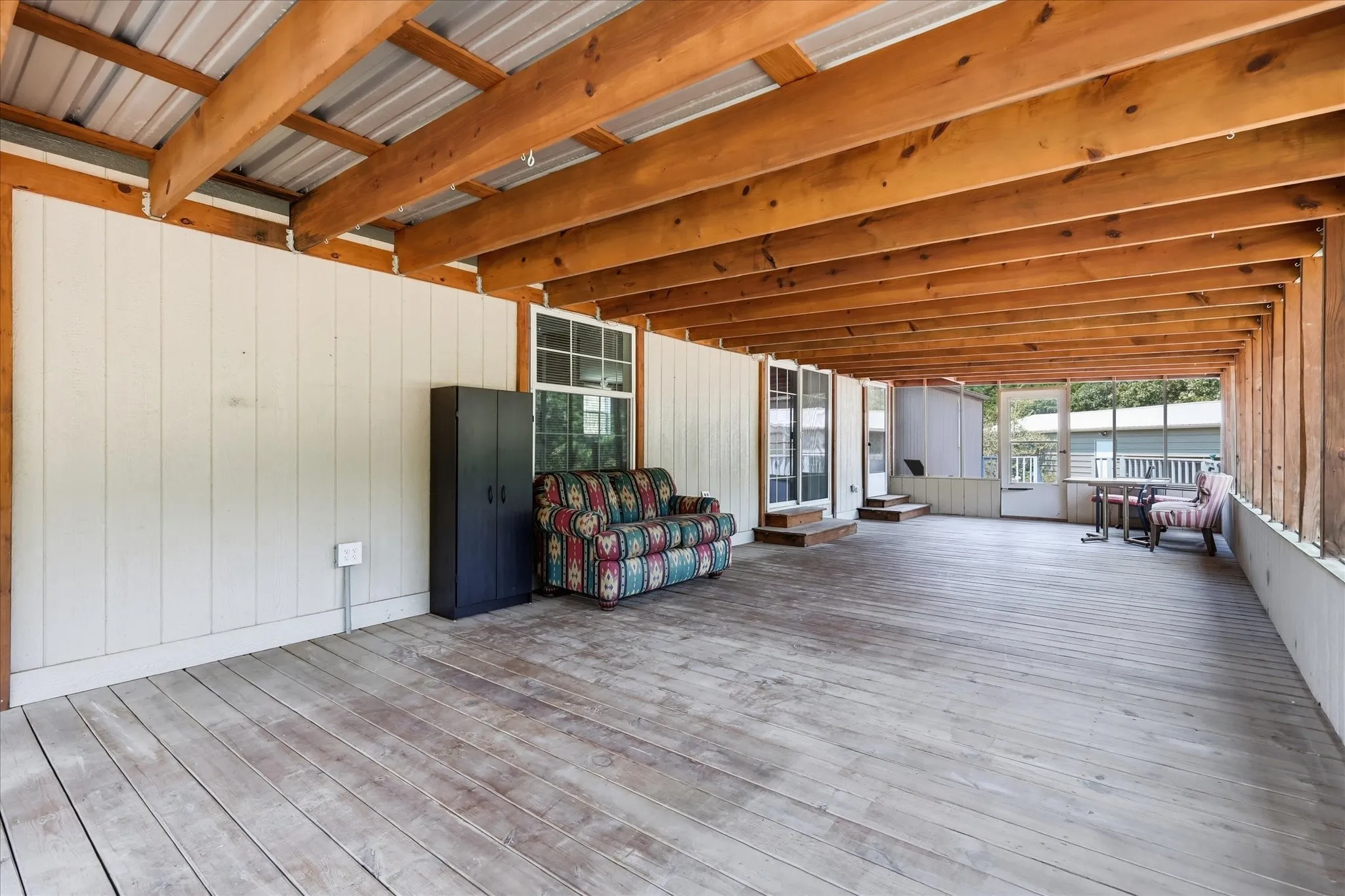 Expansive covered outdoor living space featuring a wood-finish deck, exposed timber ceiling beams, and a corrugated metal roof