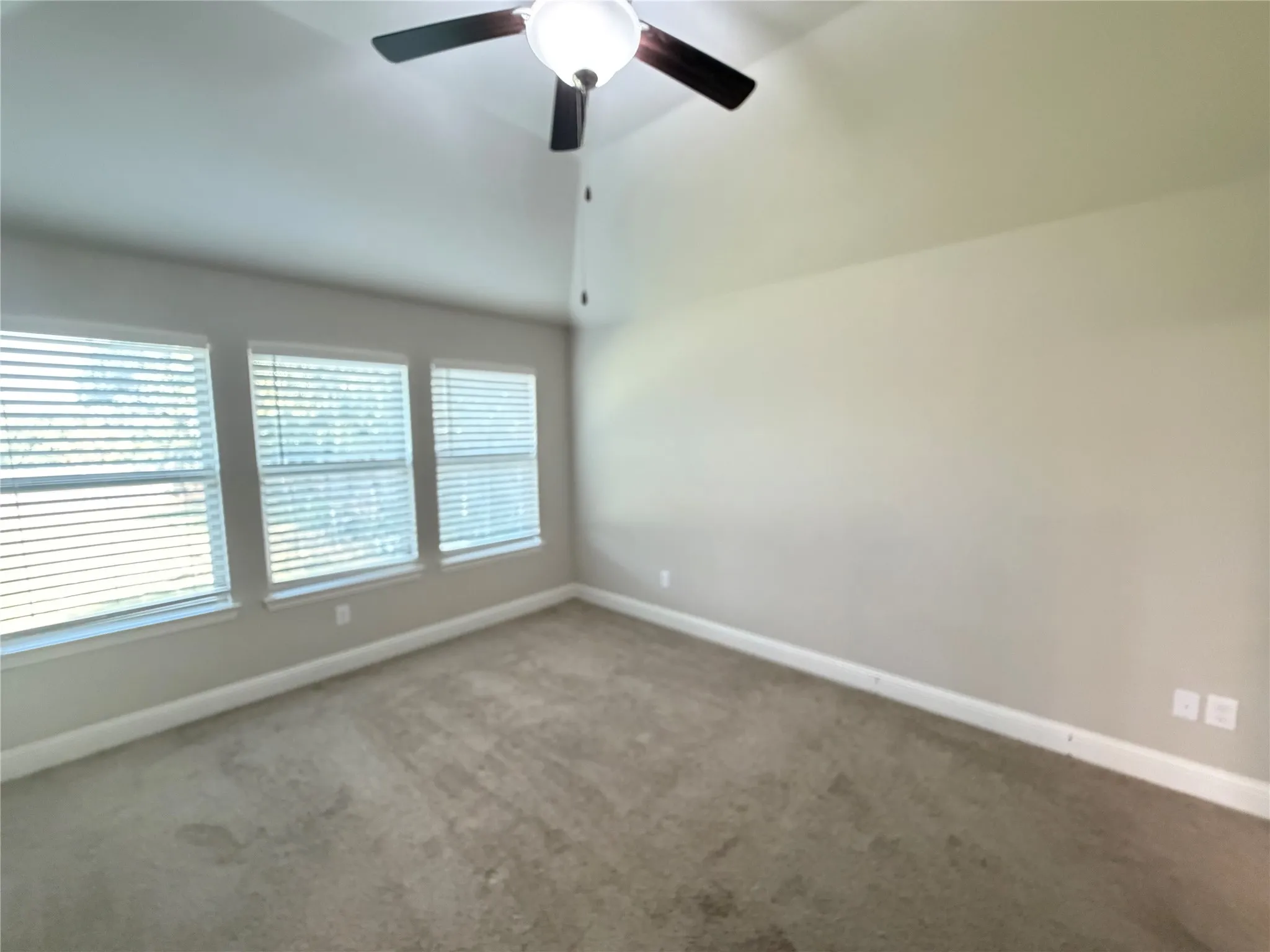 Vaulted ceiling room featuring three windows with blinds, light tan wall paint, and neutral carpet flooring