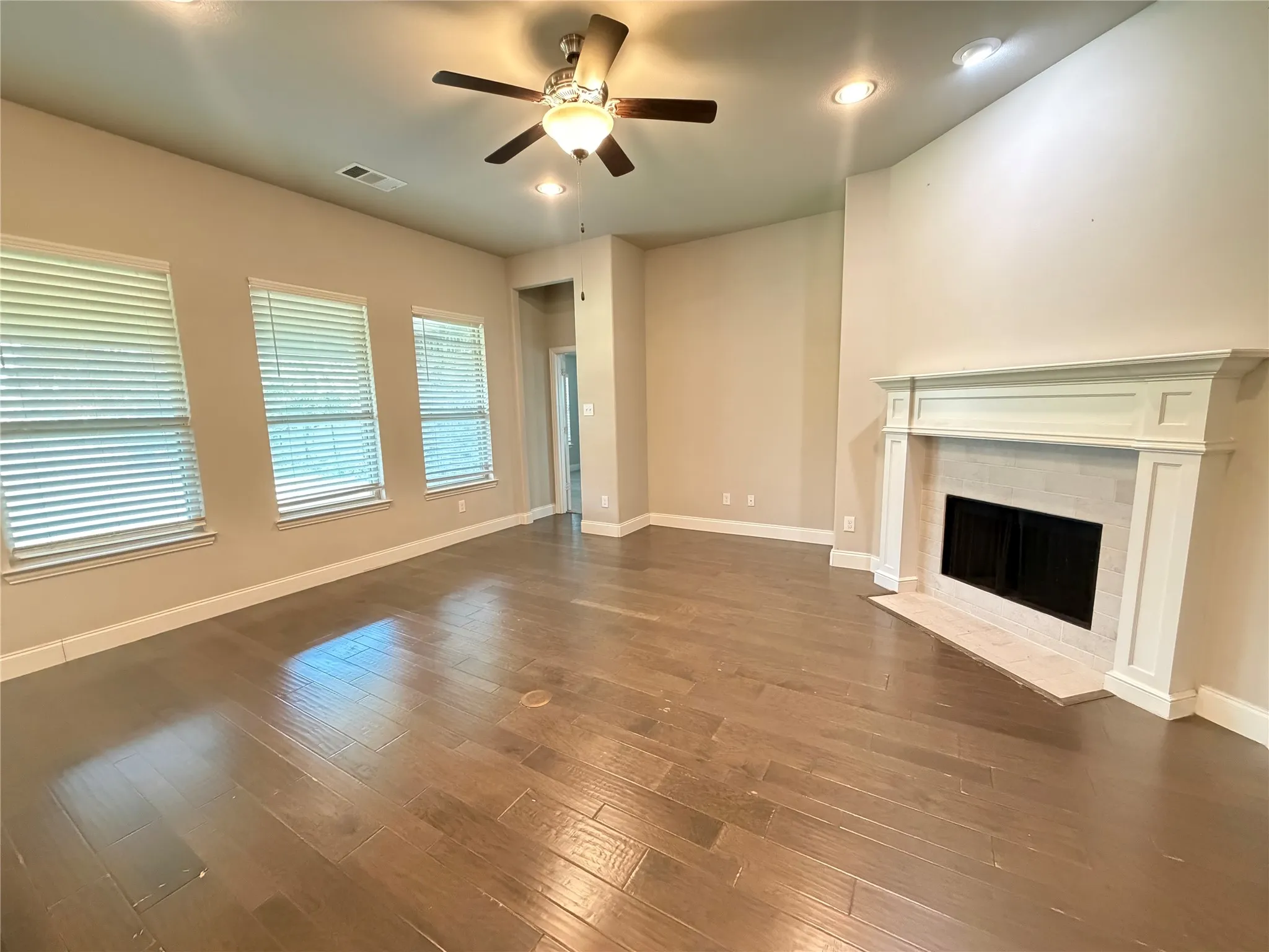 Spacious room featuring wood-finish flooring, a decorative fireplace with a white mantel and tile surround, three windows with blinds, recessed lighting, and a ceiling fan