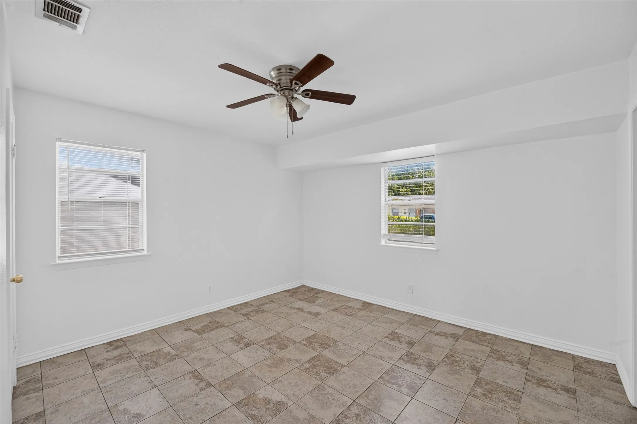 Spacious room featuring neutral-toned tile flooring, two windows with blinds, a ceiling fan with integrated lighting, and white baseboards