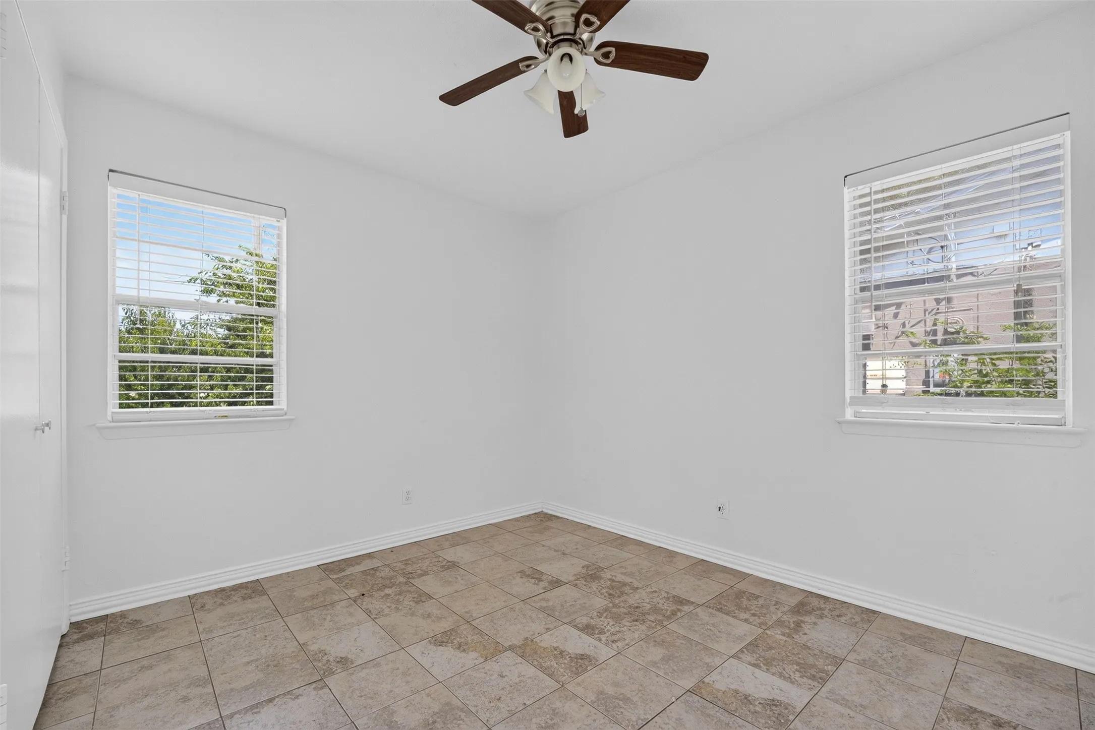 Bright interior space featuring two windows with white blinds, a ceiling fan with light fixture, white walls, and neutral-toned tile flooring