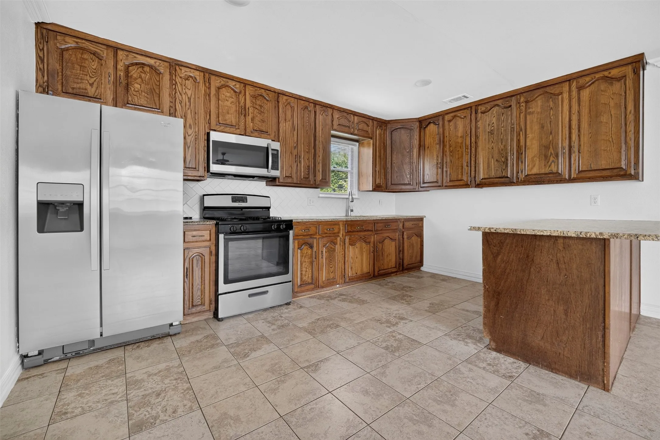 Spacious kitchen featuring warm wood cabinetry throughout, stainless steel appliances including a side-by-side refrigerator and built-in microwave, tile flooring, and a large island with a contrasting wood-finish base