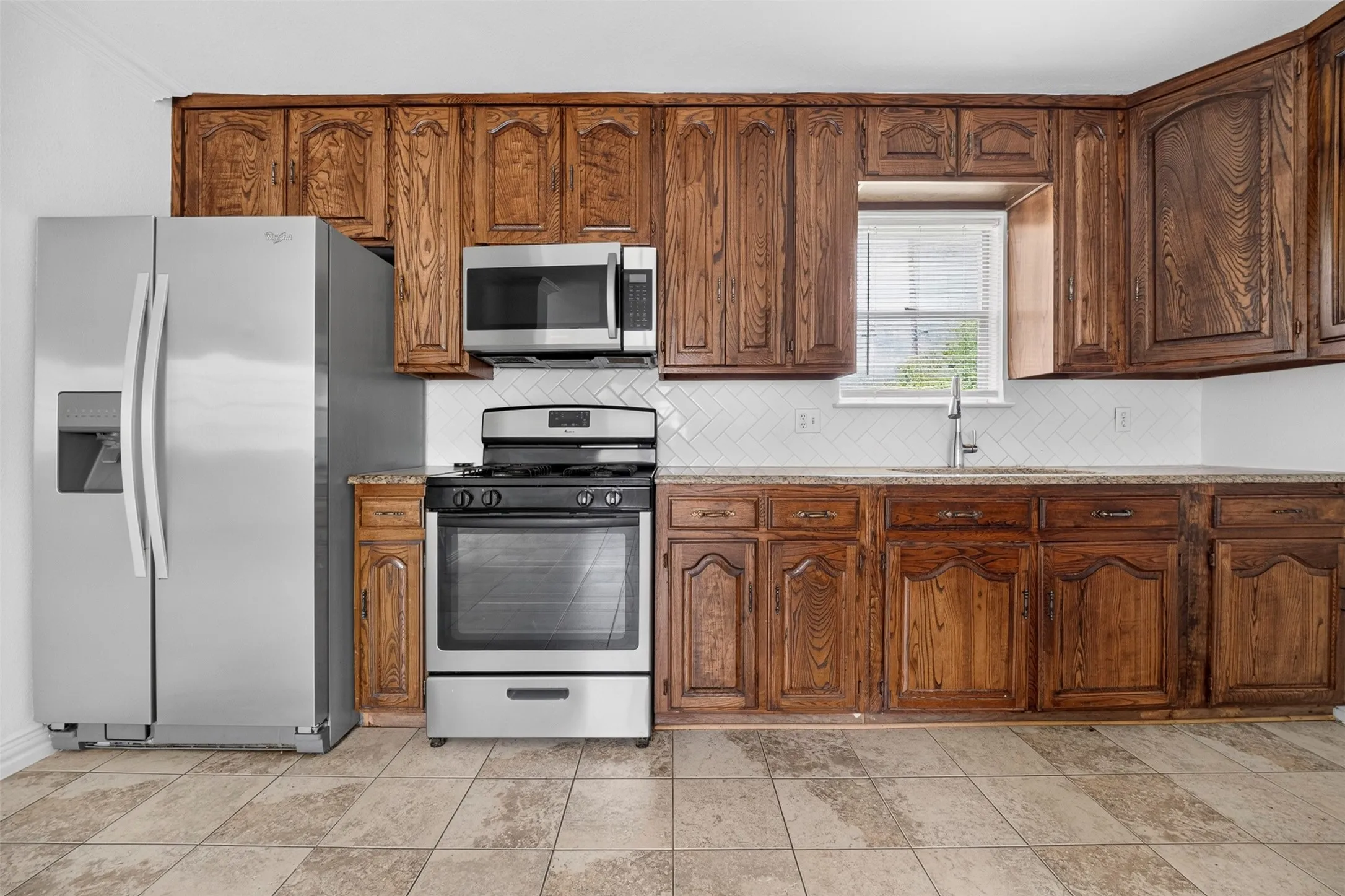 Kitchen with wood cabinetry, stainless steel appliances, granite-style countertops, white subway tile backsplash, and tile flooring