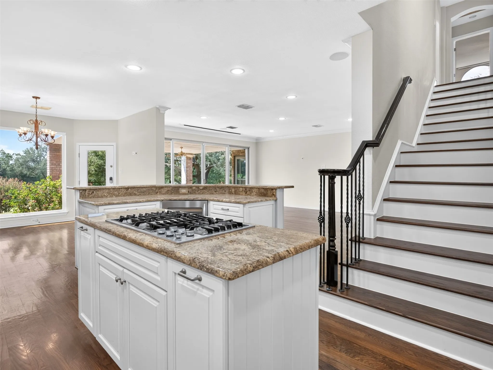 Kitchen island with integrated gas cooktop and light-toned countertop, white cabinetry, dark wood-finish flooring, and a staircase with dark wood treads and an ornate metal railing