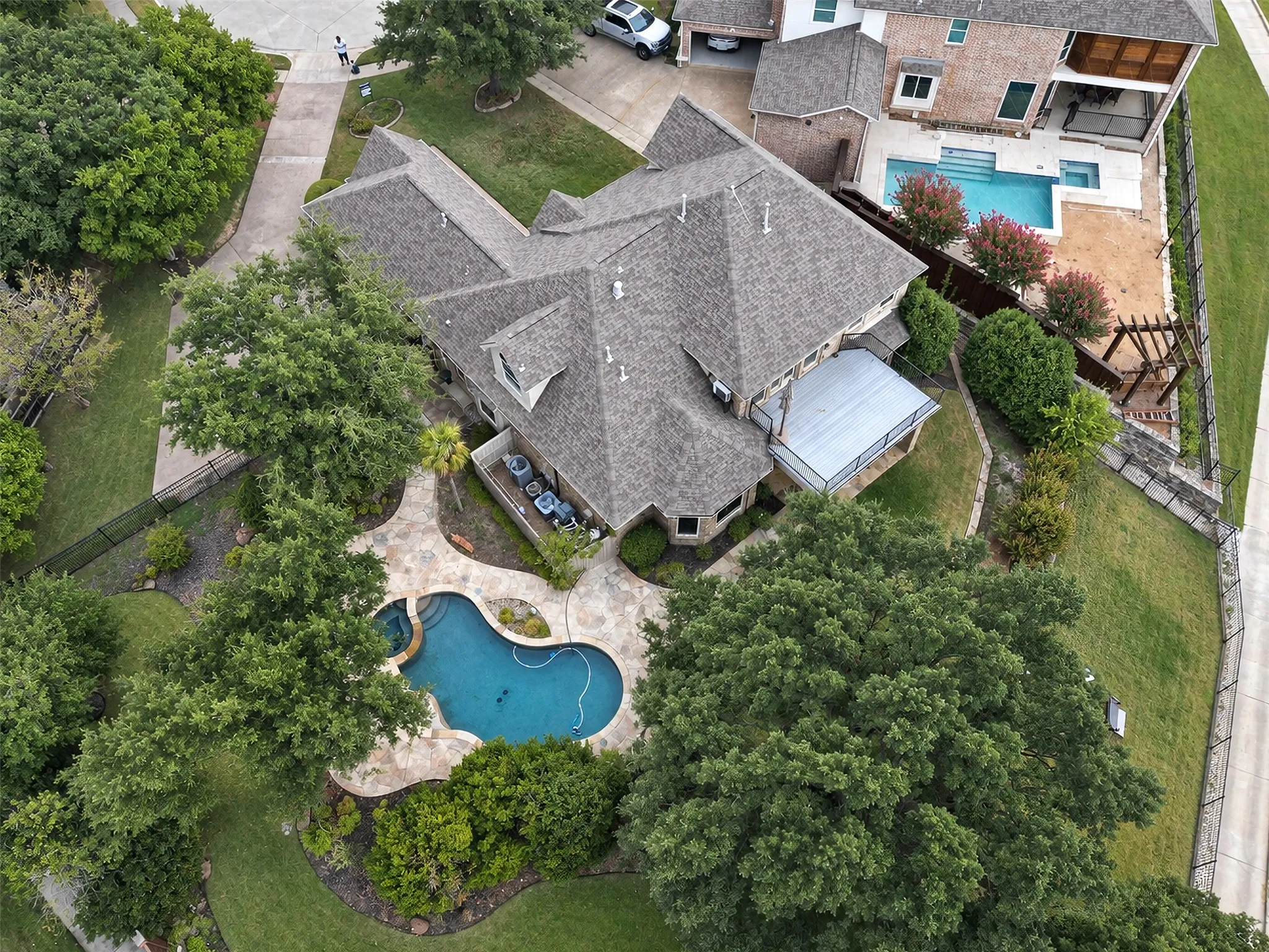 Curved stone patio with a freeform swimming pool, extensive green landscaping, mature trees, and a spacious covered patio