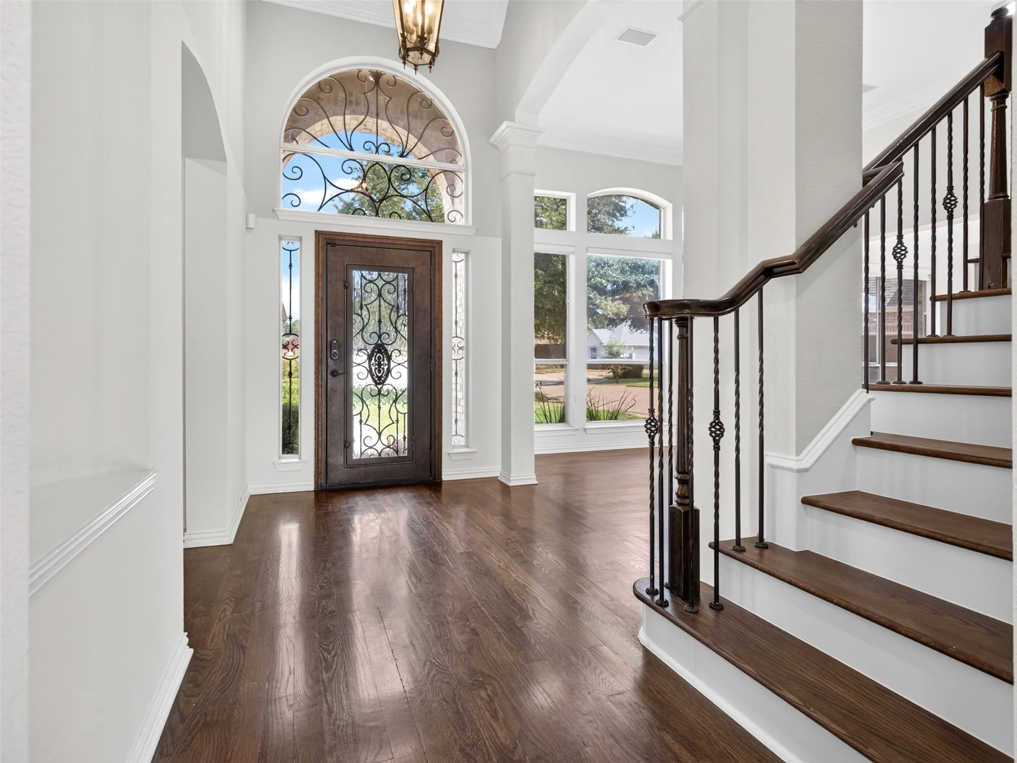 Foyer featuring wood-finish flooring, a grand arched entryway with an ornate wrought-iron door, a matching arched transom window, and a staircase with decorative metal balusters