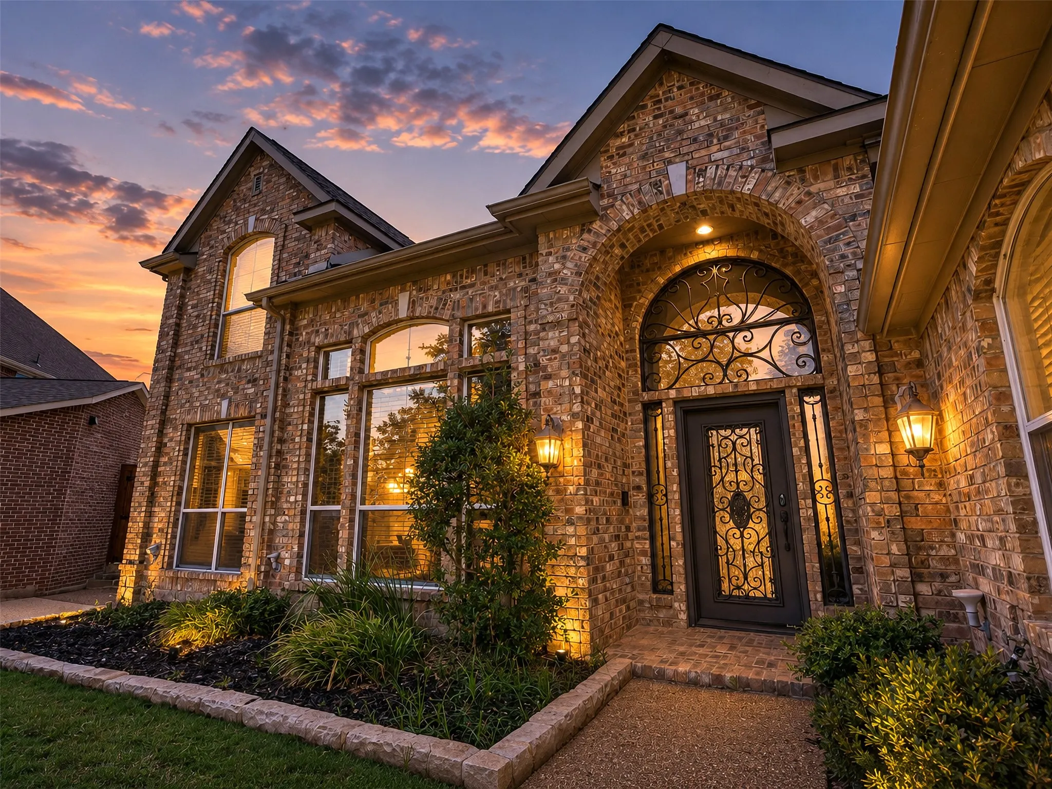 Brick exterior with arched entry, wrought iron door detailing, and landscape lighting
