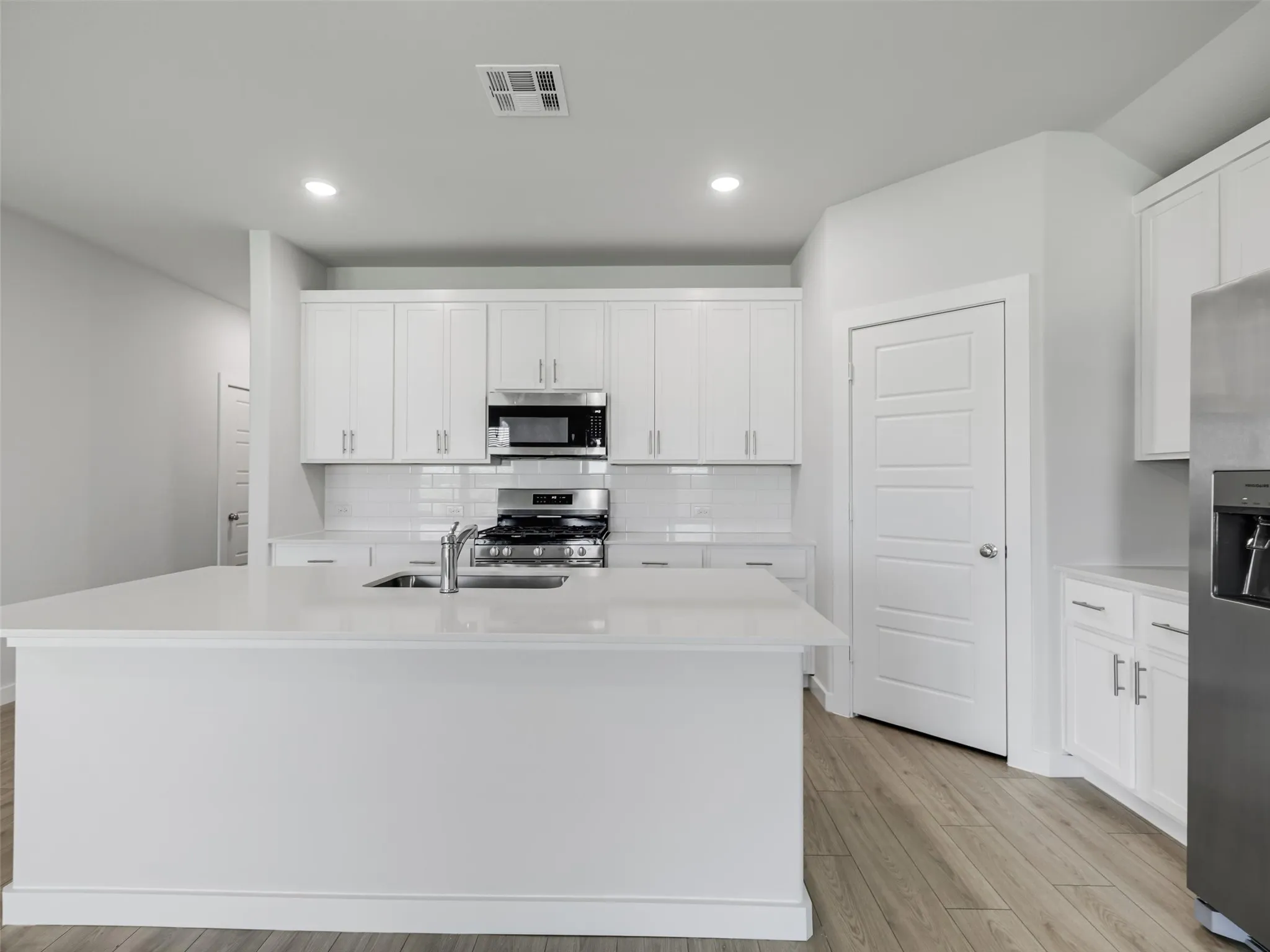 Modern kitchen featuring an expansive island with an undermount sink, white shaker cabinetry, stainless steel appliances, a subway tile backsplash, and wood-finish flooring
