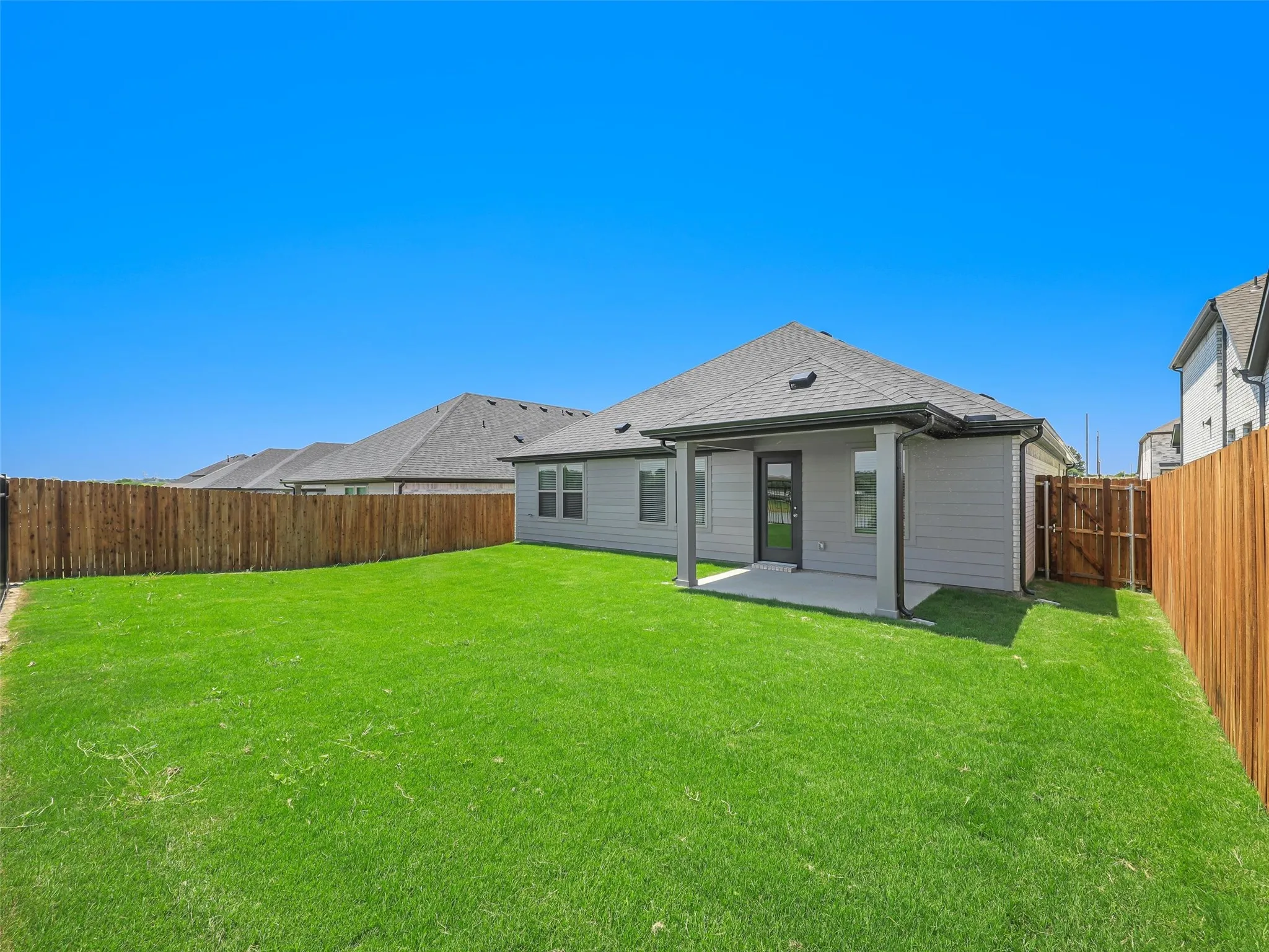 Spacious backyard featuring a manicured lawn with mature landscaping, wooden privacy fencing on both sides, and a single-story residence with a covered patio area, pitched roofline, and neutral exterior finishes