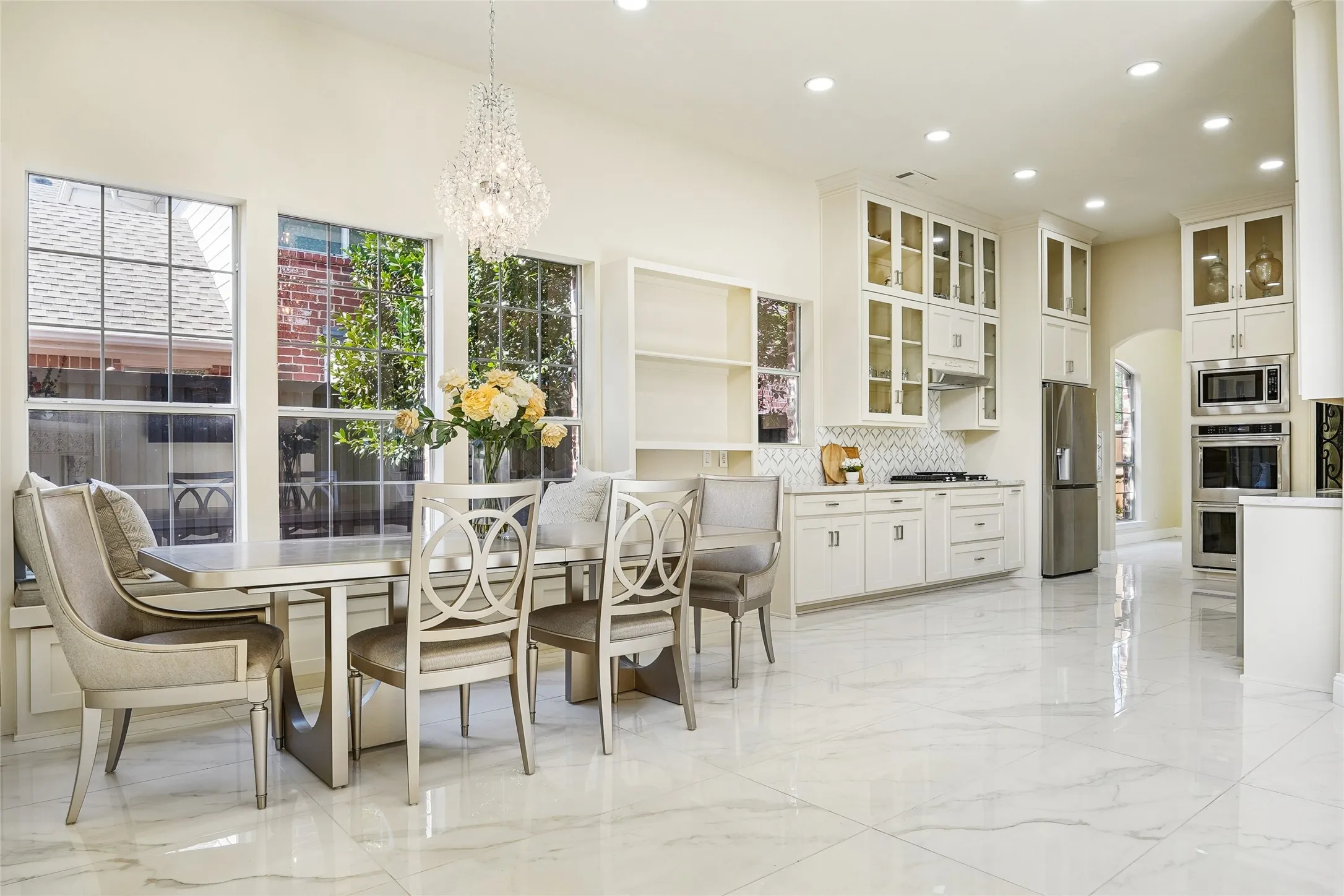 Open-concept dining and kitchen space featuring large windows, a crystal chandelier, white cabinetry with glass inserts, stainless steel appliances, and large format tile flooring