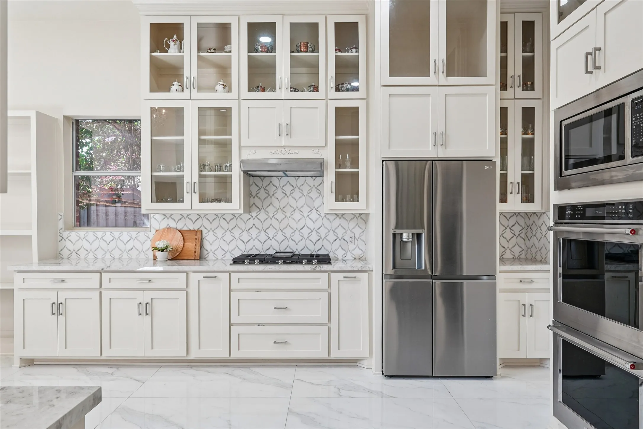 Sophisticated chef's kitchen featuring white shaker cabinetry with glass-front upper storage, honed marble countertops, and a geometric tile backsplash