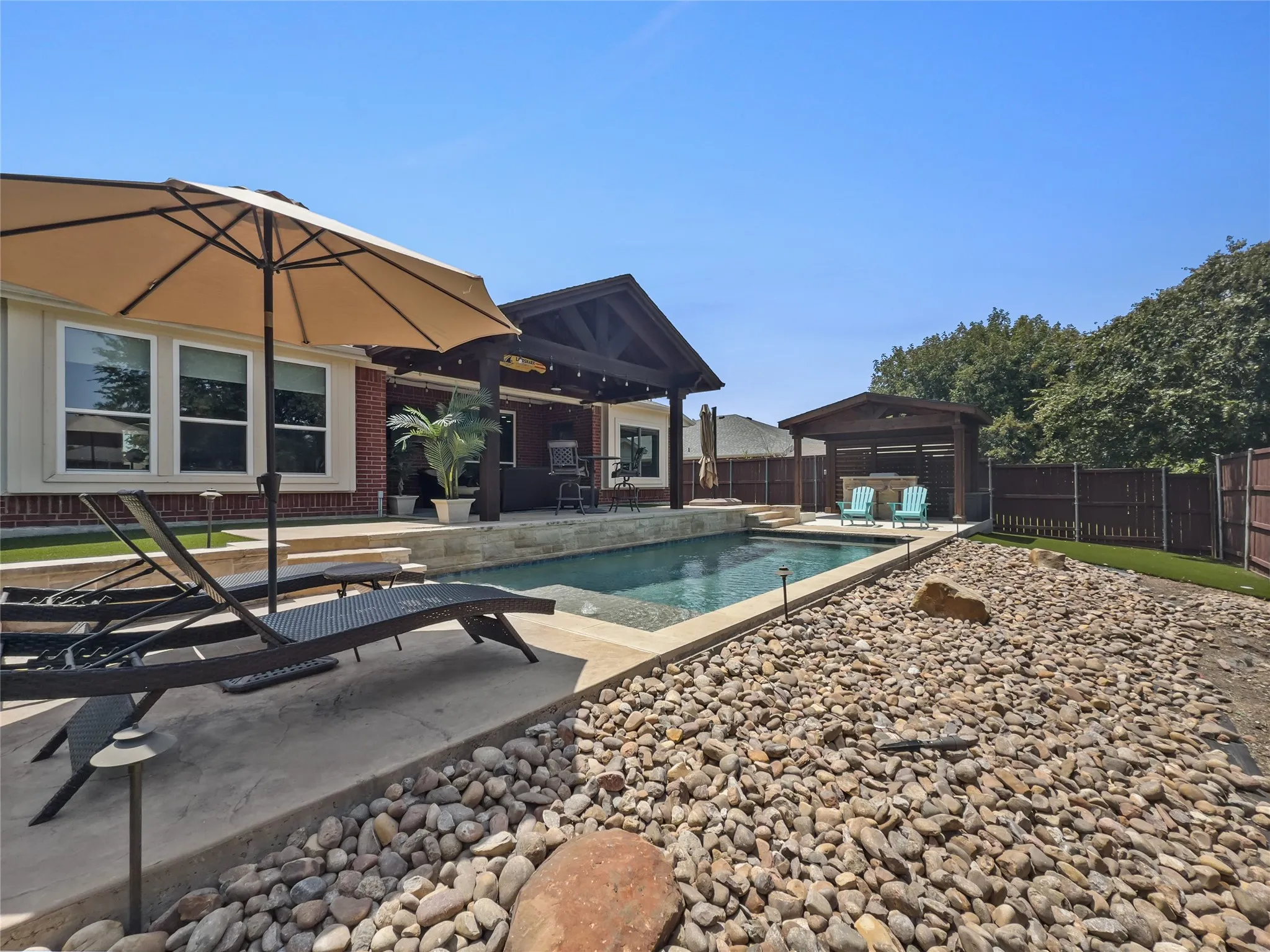 Backyard featuring a built-in swimming pool, covered patio, outdoor kitchen structure, and rock landscaping