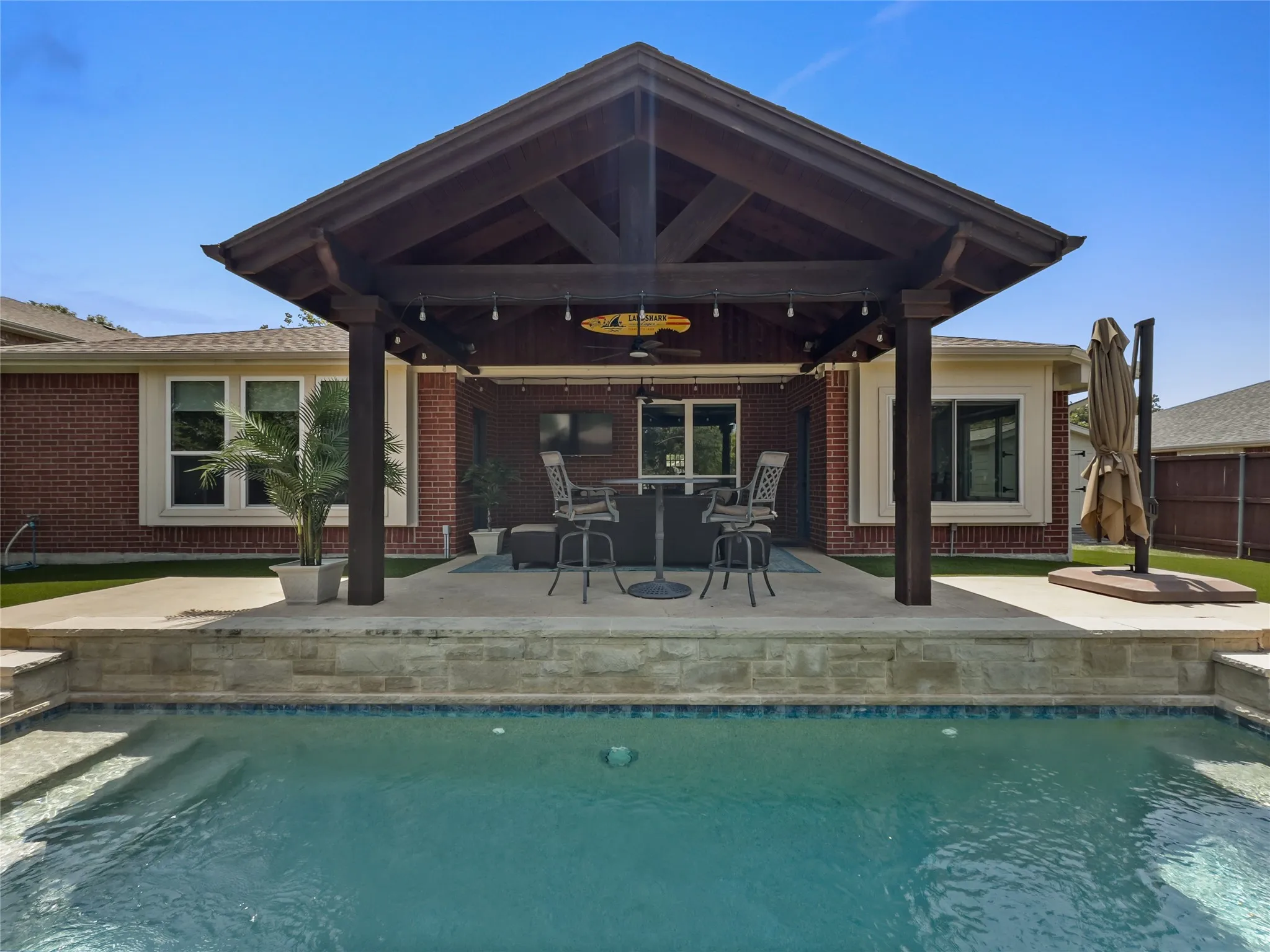 Outdoor living space featuring a swimming pool, a covered patio with exposed beams, brick exterior, and a stone-clad retaining wall