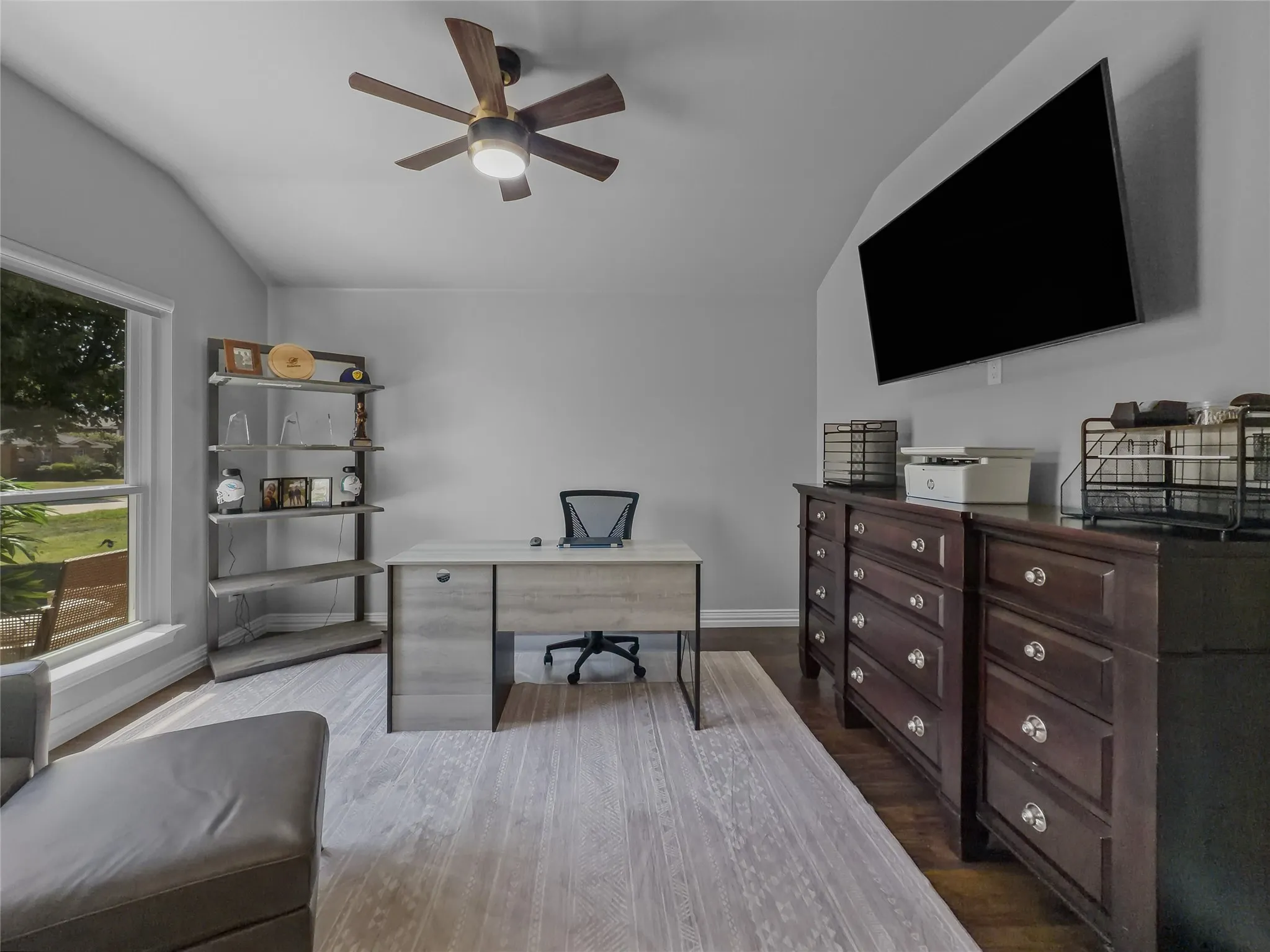 Vaulted ceiling with a contemporary ceiling fan, large window with natural light, wood-finish flooring, and wall-mounted display