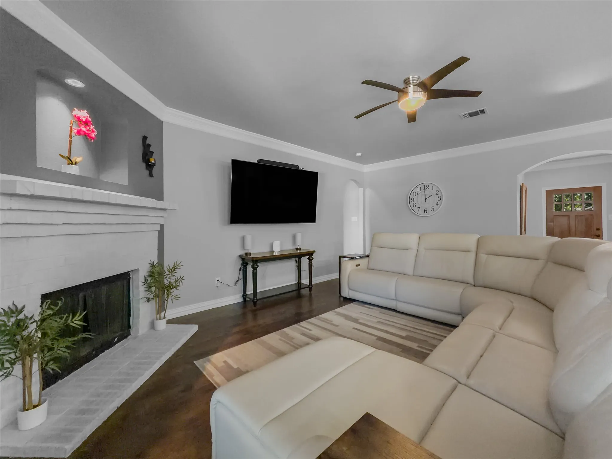 Painted brick fireplace with hearth, built-in recessed wall niches, crown molding, wood-finish flooring, and a ceiling fan with integrated lighting