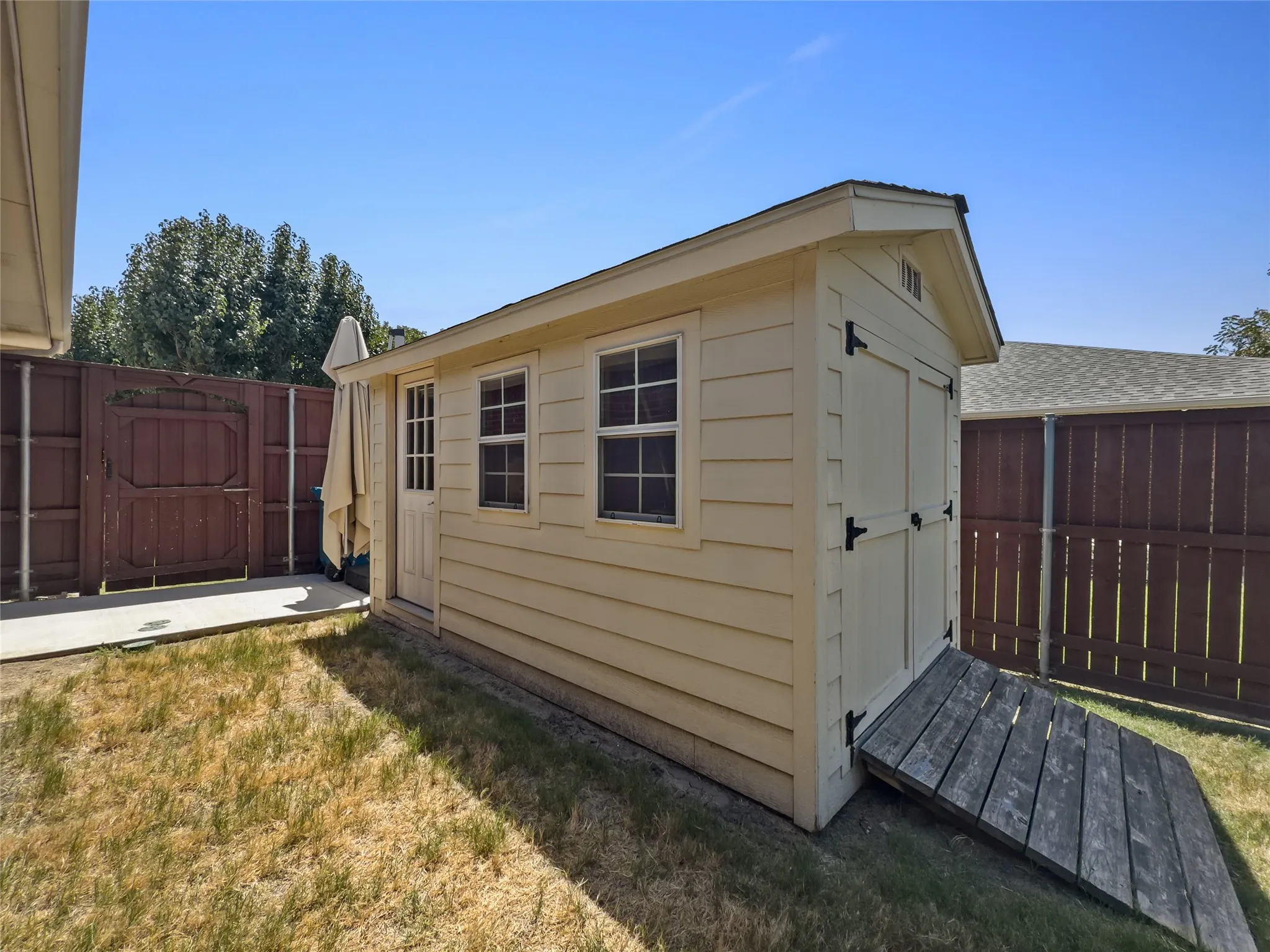 Charming storage shed featuring horizontal siding in a neutral cream finish, double-hung windows with black hardware, a pitched roof with asphalt shingles, and a wooden ramp entry