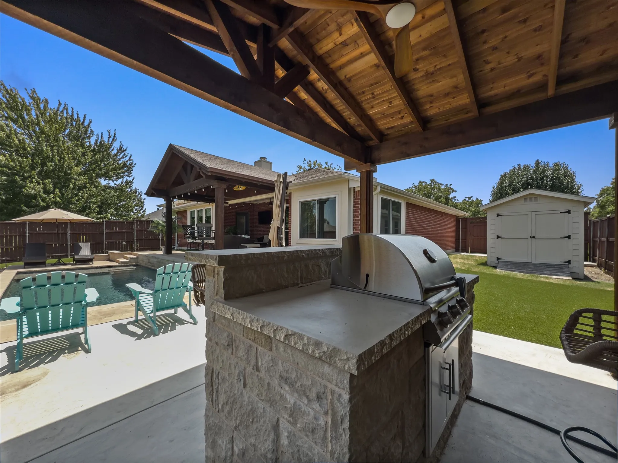 Outdoor kitchen with stone veneer base and built-in grill, covered patio with wood ceiling, swimming pool, and detached storage shed