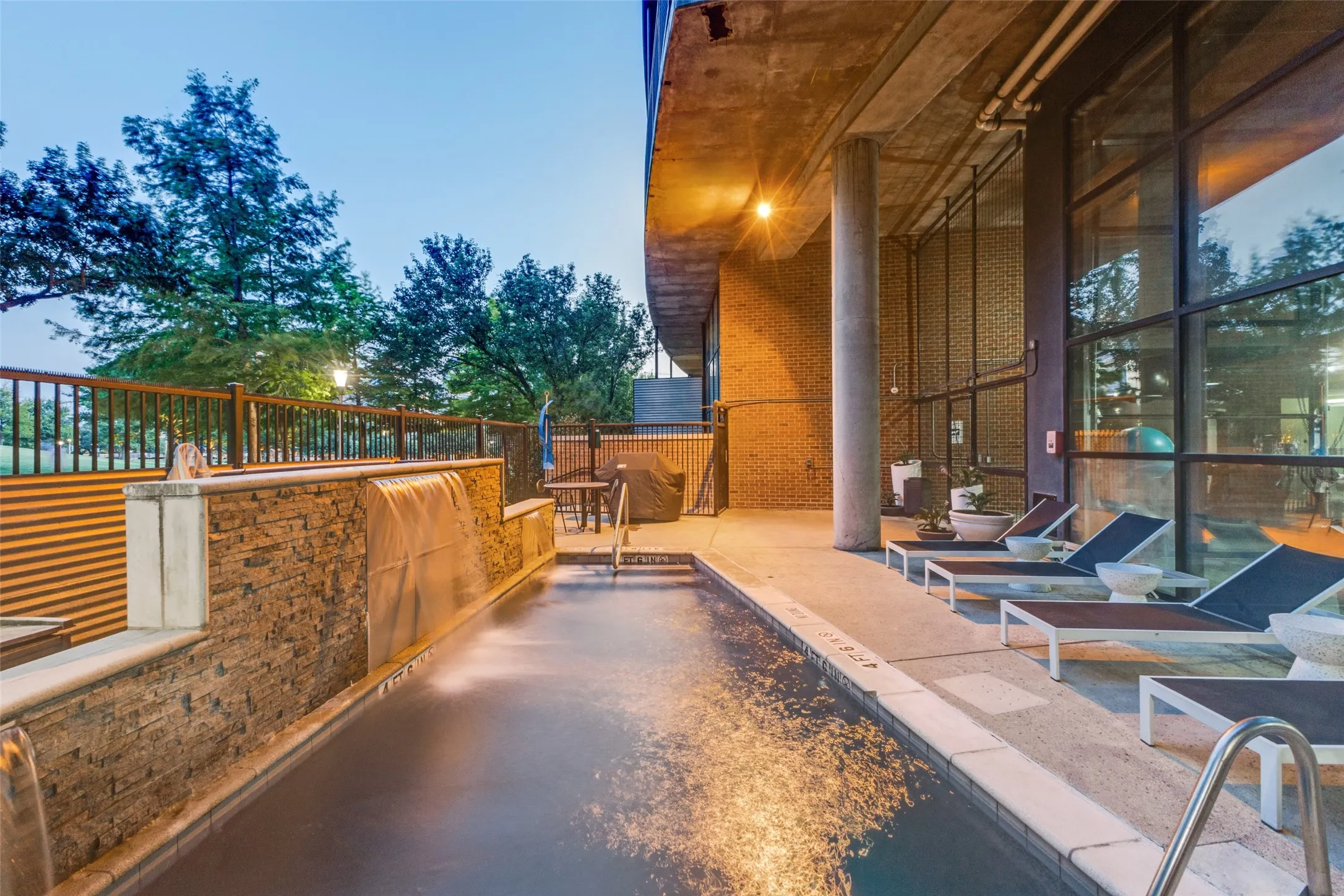 Swimming pool with integrated waterfall feature, stone accent wall, concrete decking, and glass-paneled building exterior