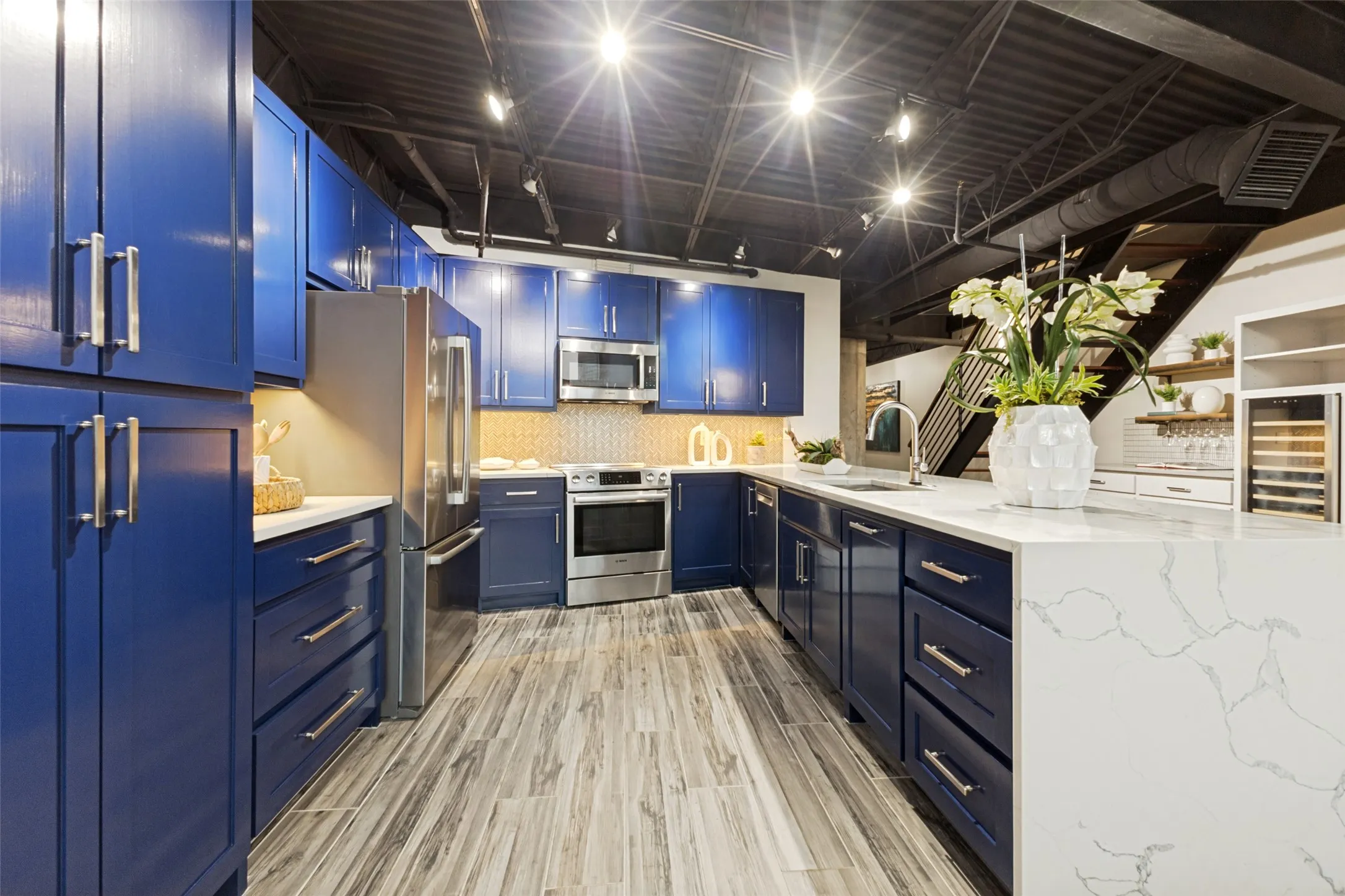 Kitchen featuring blue cabinetry, stainless steel appliances, white patterned countertops, wood-finish flooring, and exposed industrial-style ceiling elements