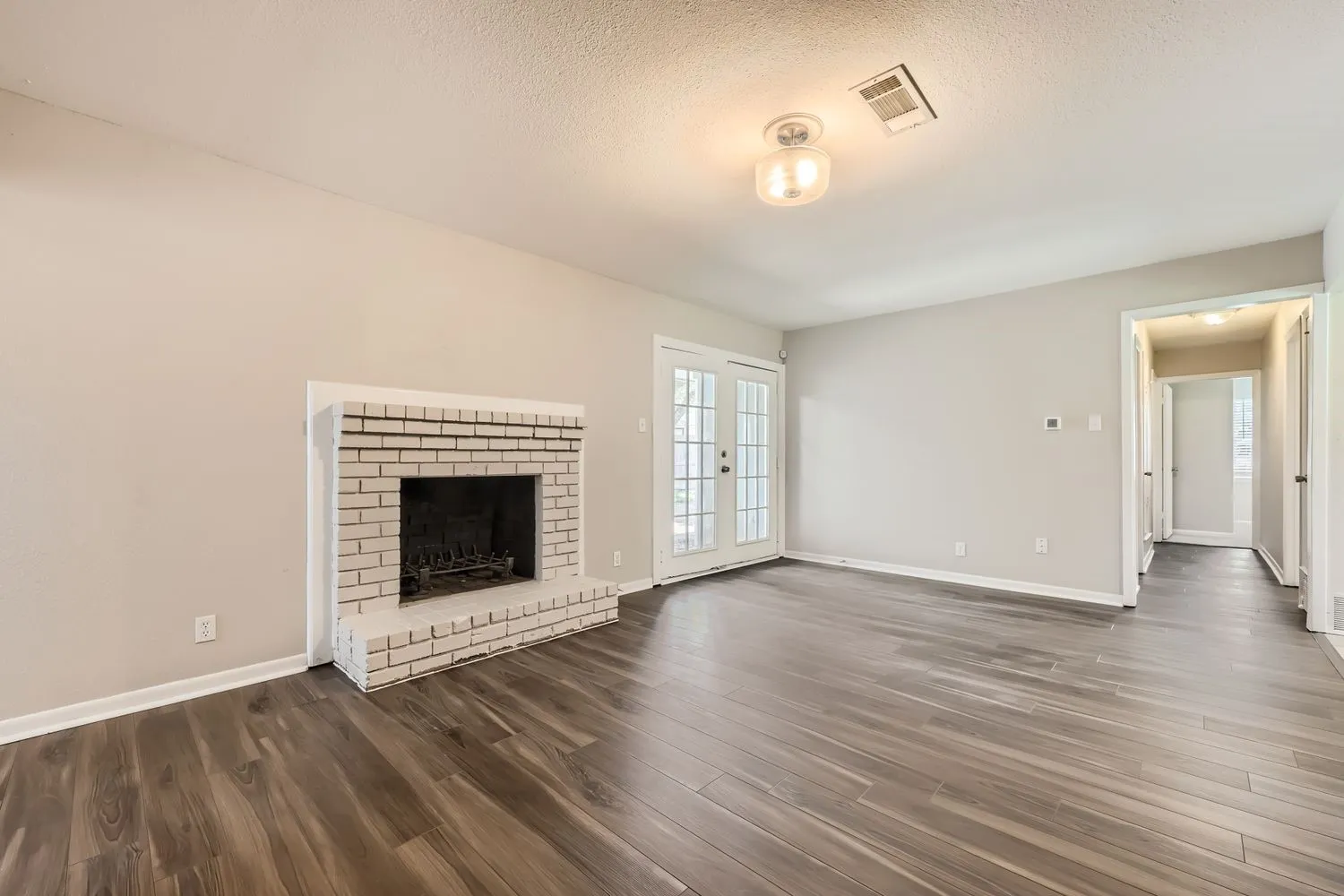 Spacious living area showcasing gleaming wood-finish flooring throughout, a striking brick fireplace with dark surround, and abundant natural light from French doors