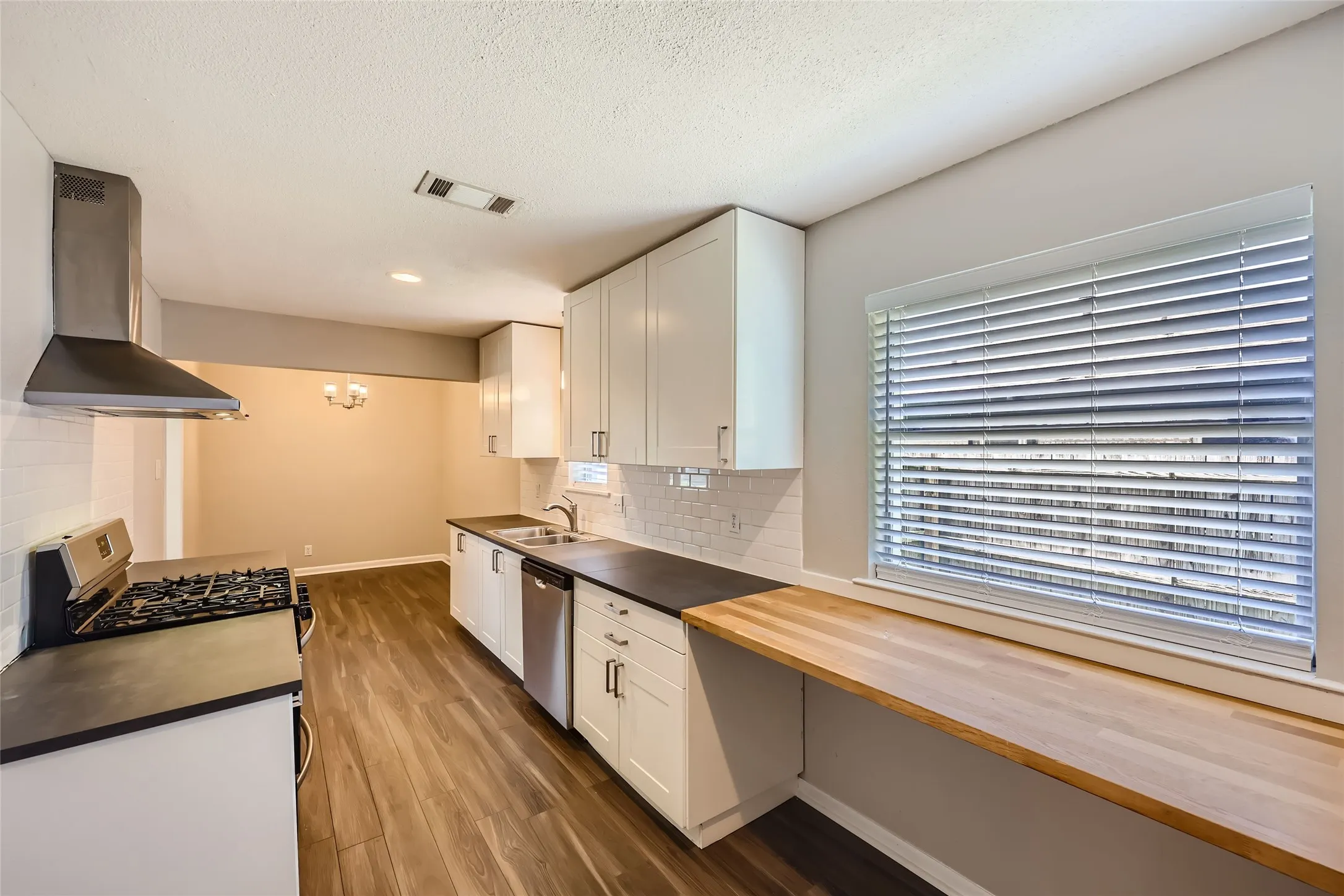 Modern kitchen featuring white cabinetry, wood-finish countertops, stainless steel appliances including a gas cooktop and range hood, and abundant natural light from large horizontal blinds