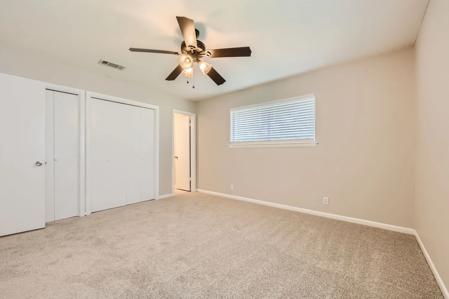 Spacious room featuring neutral carpeting, a ceiling fan with integrated lighting, window with blinds, and two sets of bi-fold closet doors