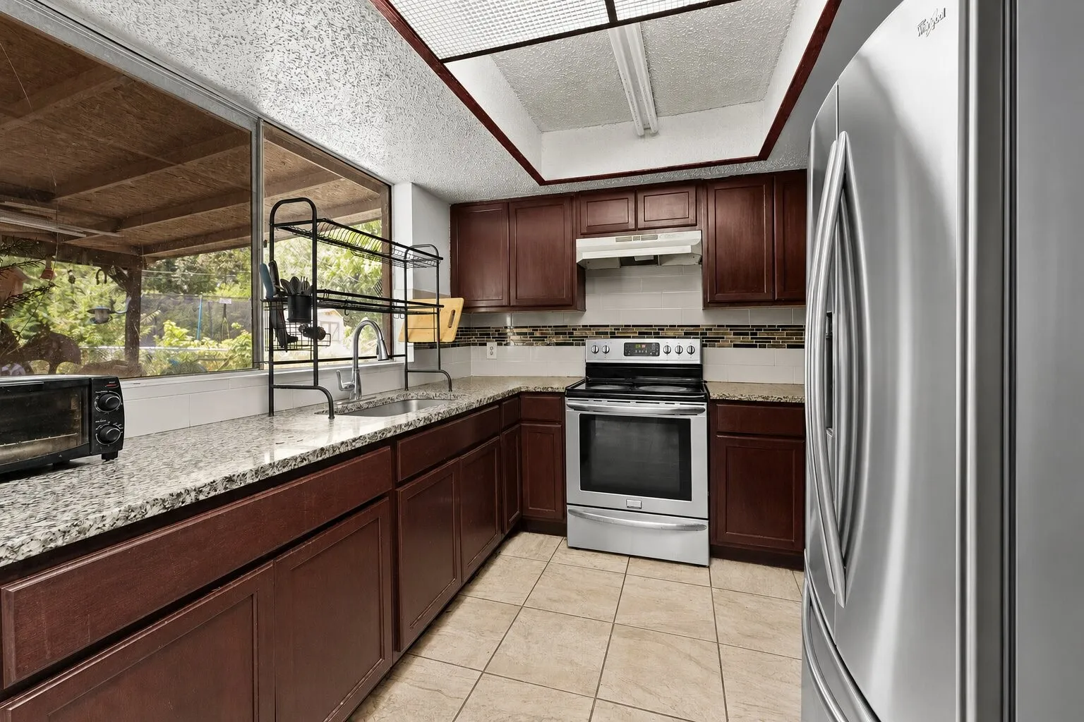 Kitchen featuring dark wood-finish cabinetry, light speckled countertops, a tile backsplash, stainless steel appliances, and a window overlooking a covered outdoor area