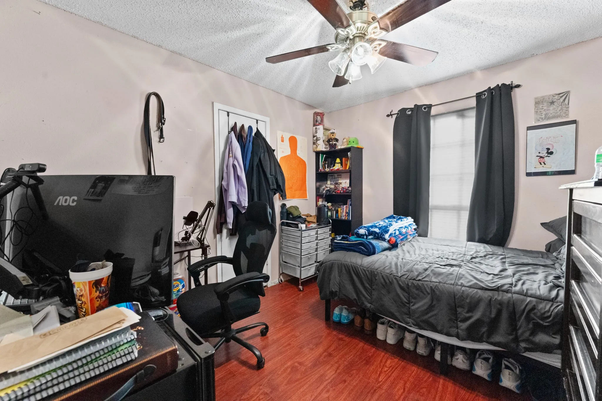 Spacious bedroom featuring wood-finish flooring, ceiling fan with integrated lighting, and ample natural light from large window with blackout curtains
