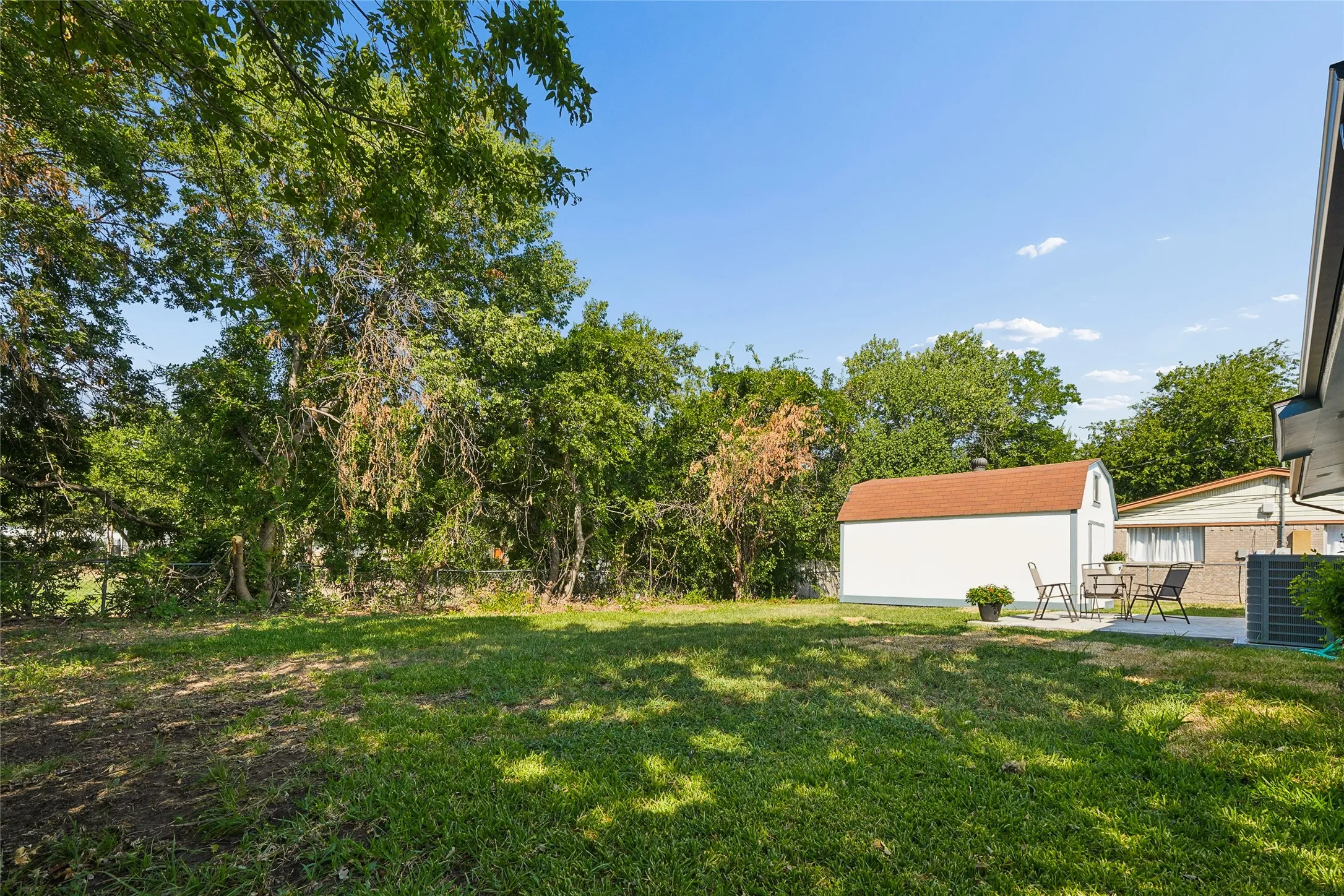 Expansive backyard featuring a manicured lawn, established trees, a concrete patio, and a white storage shed with a gambrel roof