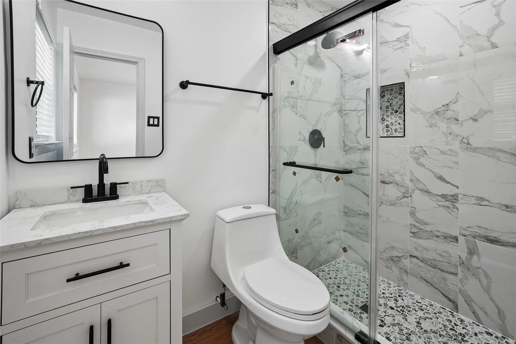Bathroom featuring a white vanity with an integrated sink and matte black fixtures, a framed mirror, and a glass-enclosed shower with large format marble-look tile walls and a pebble-style floor