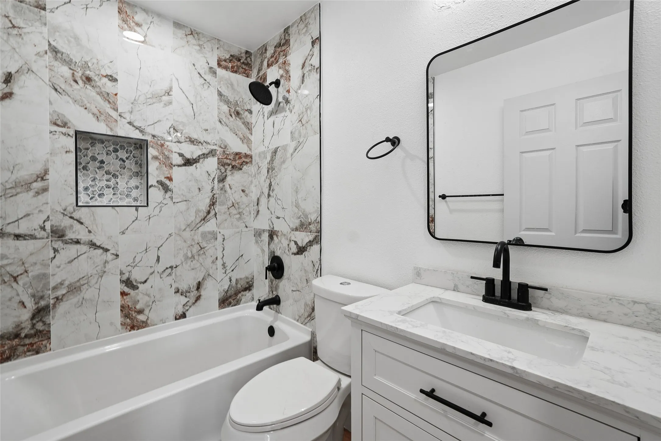 Spa-inspired bathroom featuring a marble-look tile surround with coordinating floor, white vanity with integrated sink, quartz countertop, matte black hardware and fixtures, framed mirror with metal detailing, and separate soaking tub with rainfall showerhead
