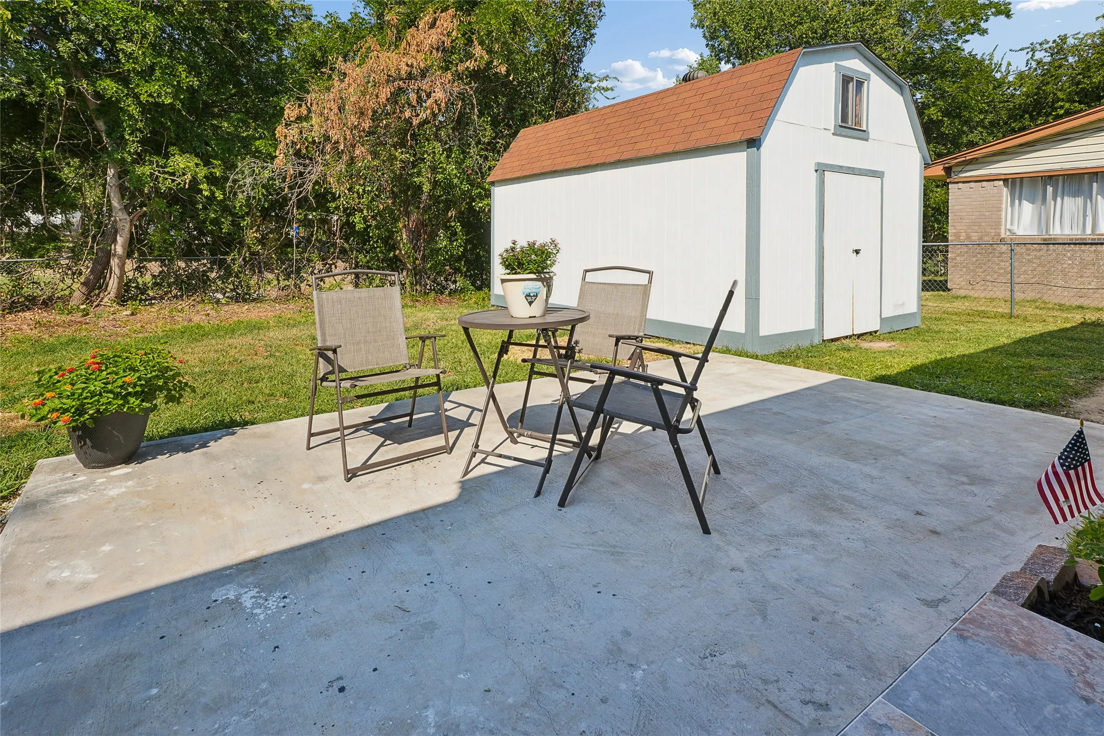 Concrete patio with a freestanding shed featuring white siding and an orange shingled roof, surrounded by a grassy yard and mature trees