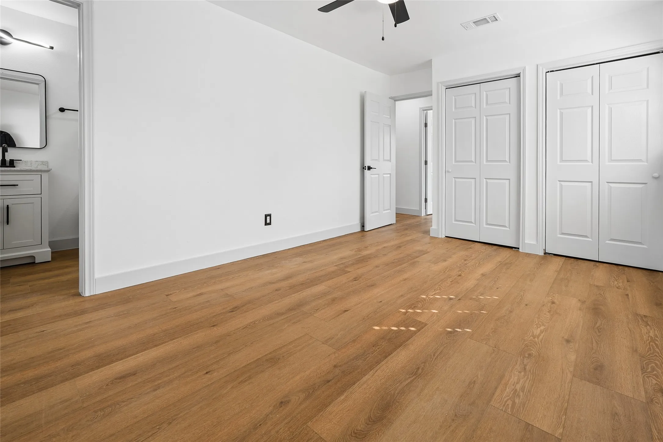 Spacious primary bedroom featuring warm wood-finish flooring, white six-panel closet doors, ceiling fan with integrated lighting, and neutral wall finishes with an electrical outlet