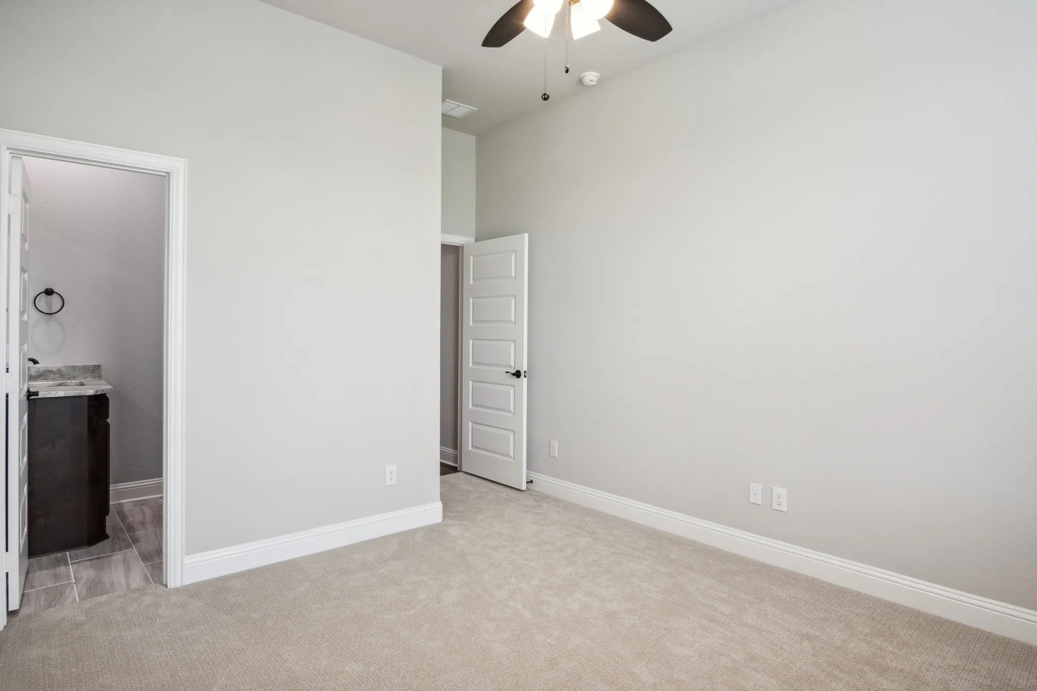 Carpeted room featuring a ceiling fan, white baseboards, and a doorway leading to a bathroom with a dark wood-finish vanity, light-colored countertop, and tile flooring