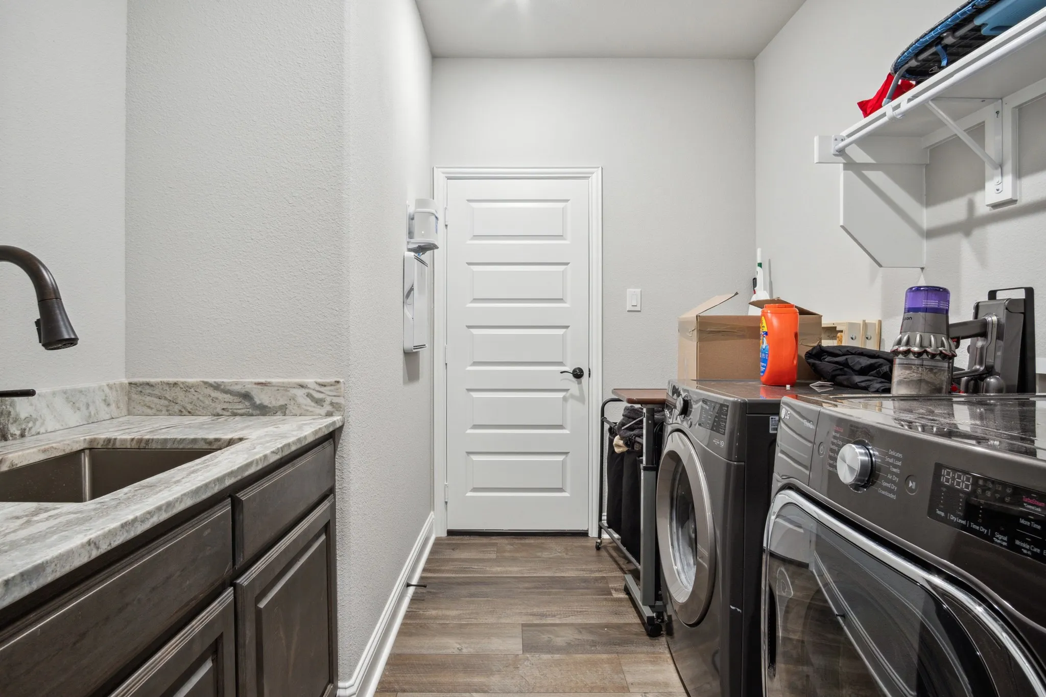 Utility room featuring wood-finish flooring, a stainless steel sink with a dark bronze faucet, a patterned countertop, dark wood-finish cabinetry, and white wall shelving
