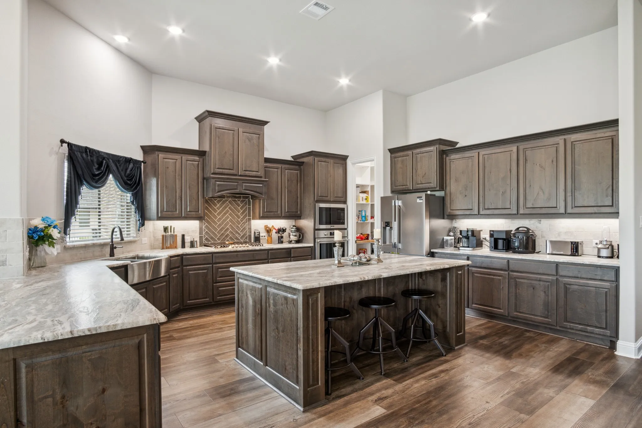 Spacious kitchen featuring dark wood-finish cabinetry, light stone countertops, a large center island with seating, a herringbone tile backsplash, and stainless steel appliances