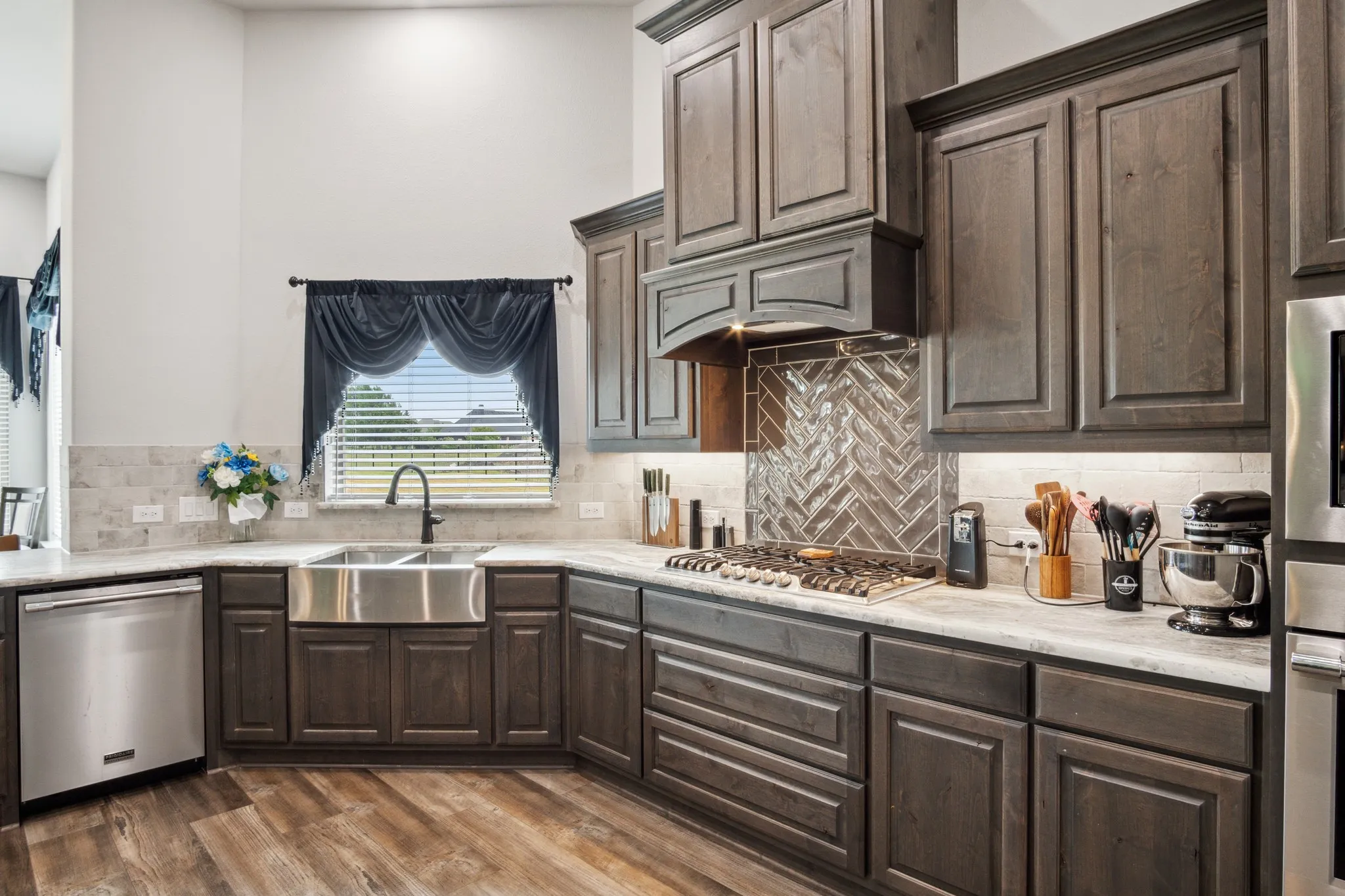 Gourmet kitchen featuring wood-finish flooring, dark wood cabinetry, stainless steel farmhouse sink, stainless steel appliances, and a herringbone pattern tile backsplash