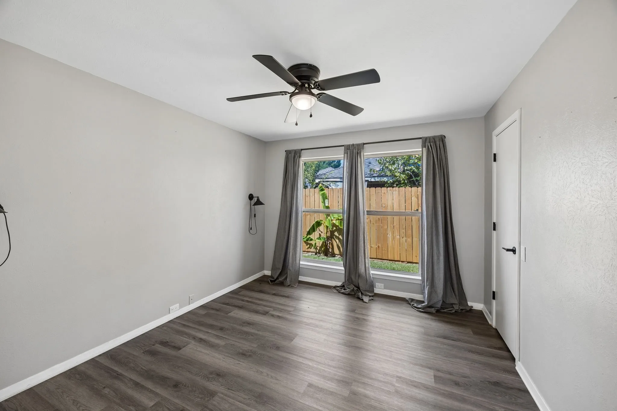Bright room featuring dual windows, gray wall paint, wood-finish flooring, a ceiling fan with integrated lighting, and a solid core door with black hardware
