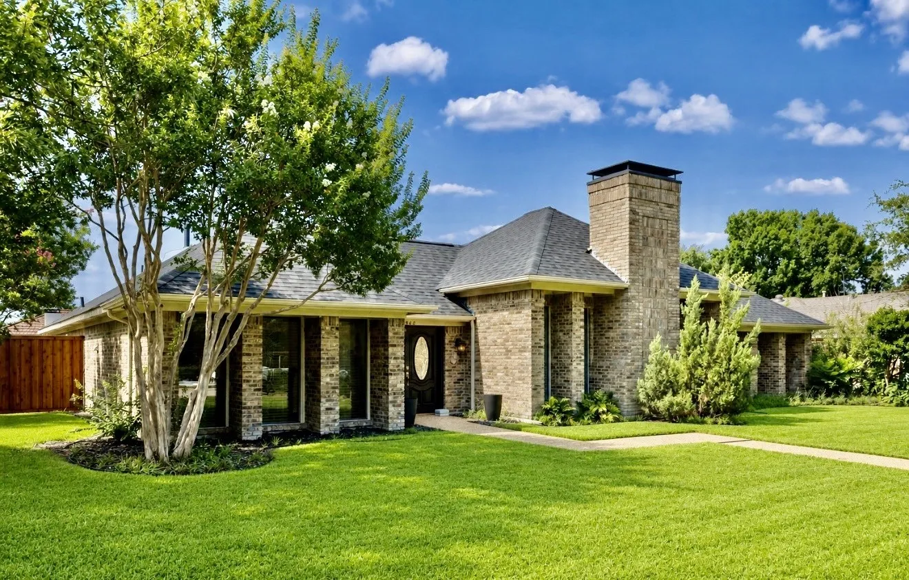 Brick exterior residence featuring a prominent chimney, dark shingle roof, and a mature tree