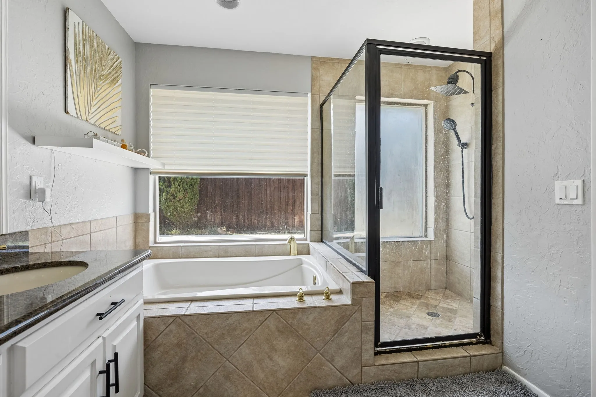 Spa-inspired primary bathroom featuring a soaking tub with tile surround, walk-in shower enclosure with black metal framing and rainfall showerhead, dual vanity with dark cabinetry, and neutral tile flooring throughout