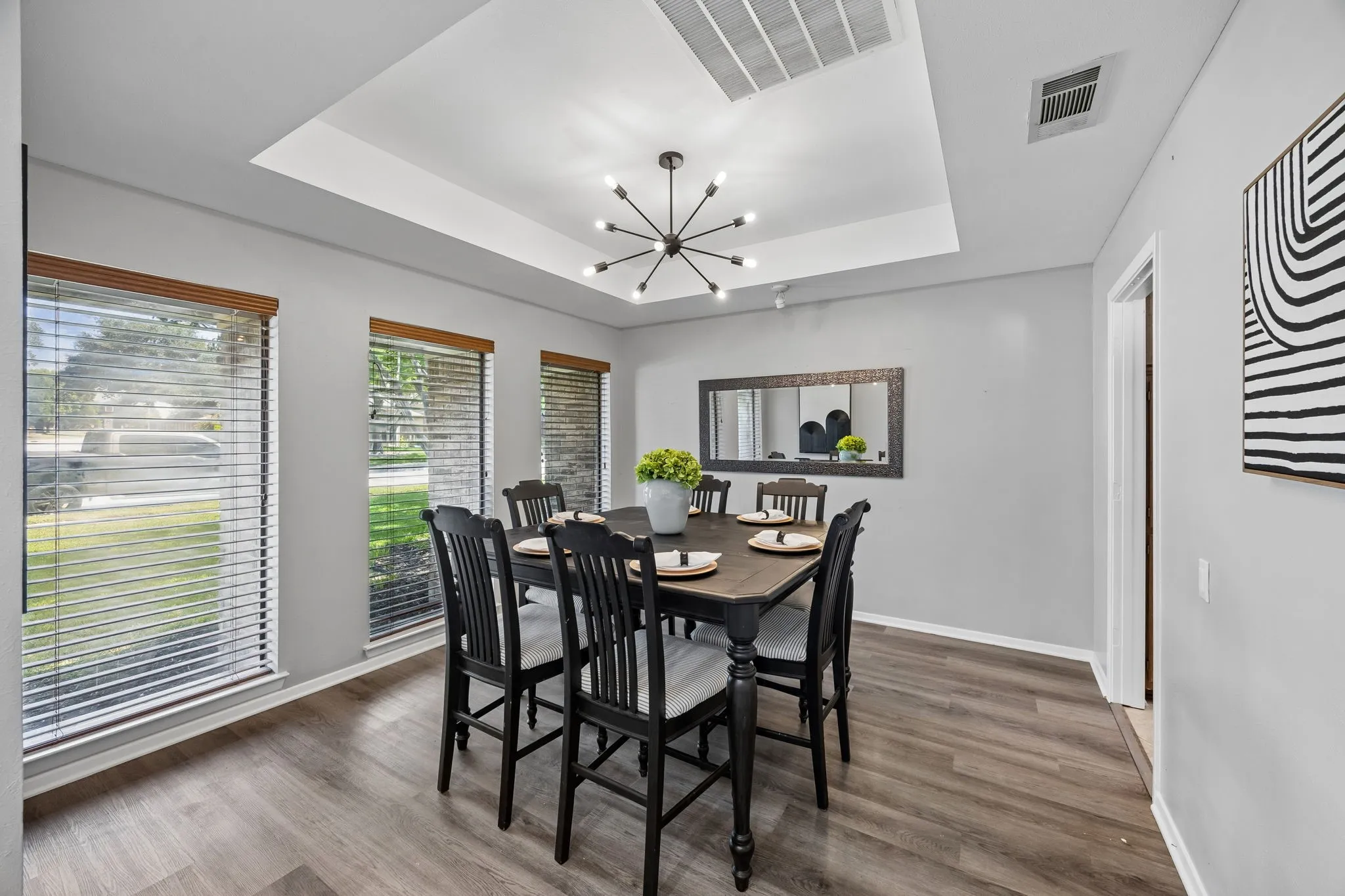Spacious dining area featuring wood-finish flooring, a tray ceiling with a modern sputnik chandelier, and multiple windows with wood-tone blinds