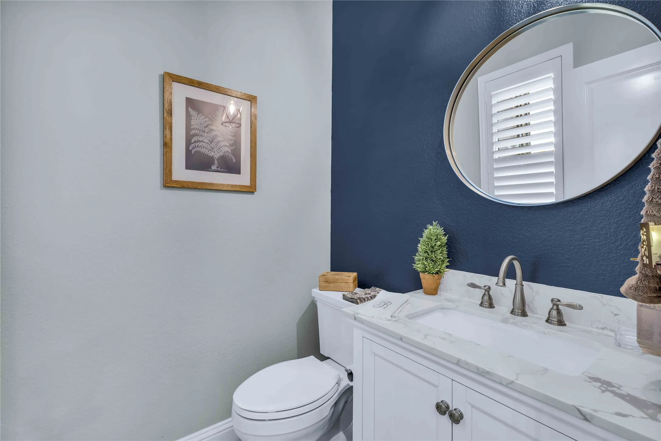 Vanity with a white countertop and undermount sink, complemented by a circular mirror, brushed nickel faucet, and white plantation shutters